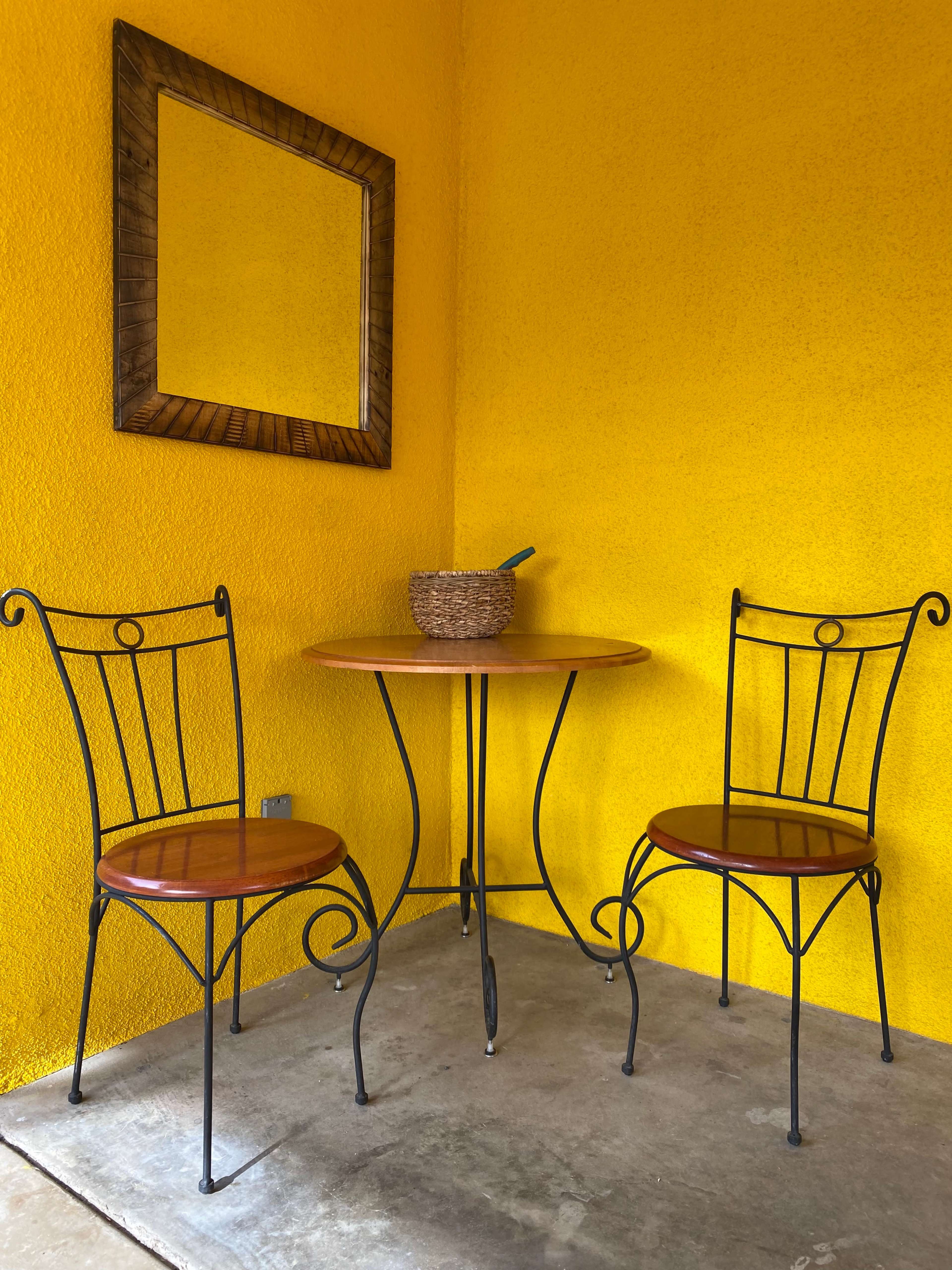 A round wooden table with two metal chairs and a woven basket sits against a bright yellow wall.
