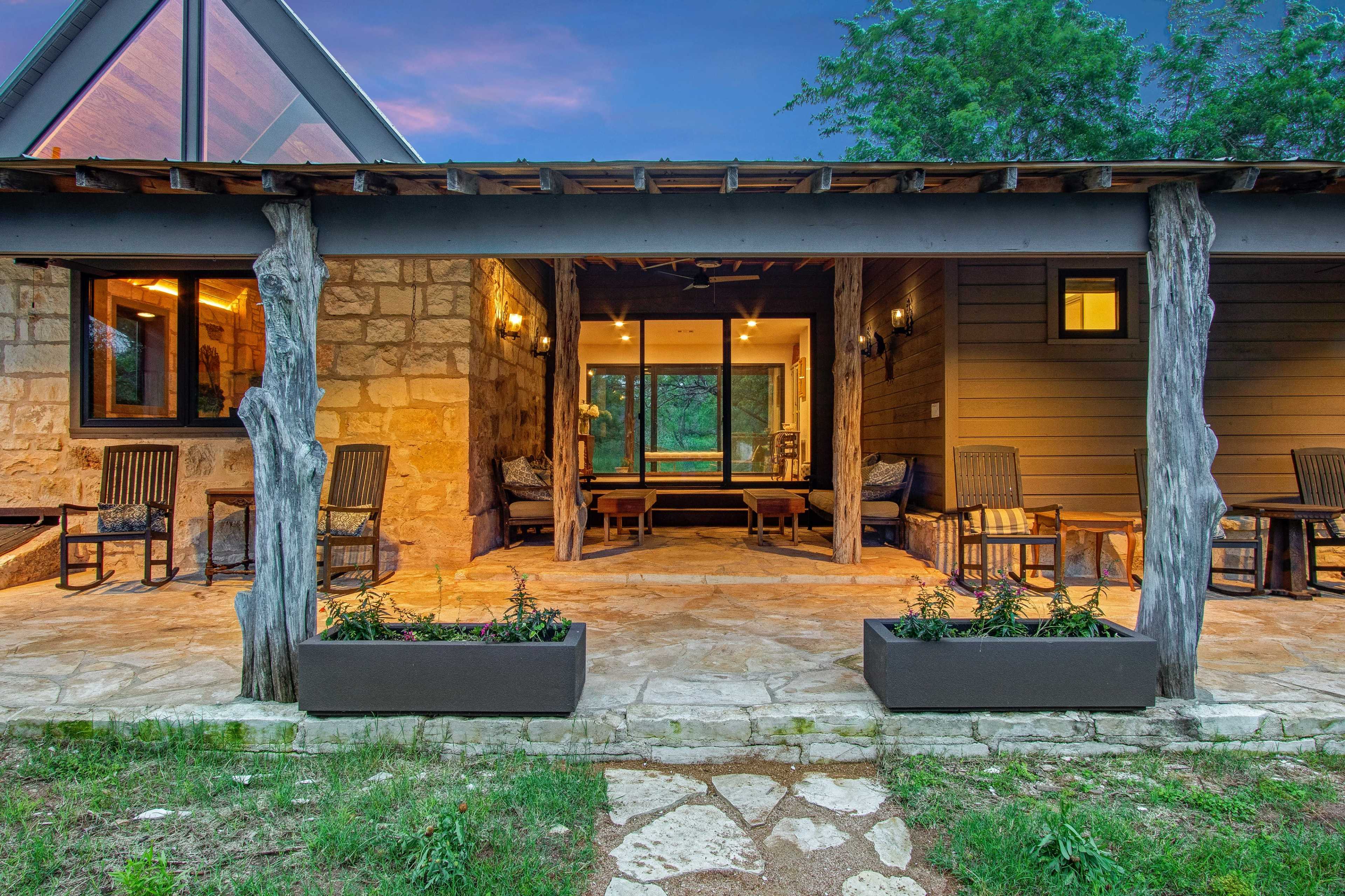 A stone and wood porch with chairs, overlooking a landscaped area leading to a sliding glass door that opens into a well-lit interior space.