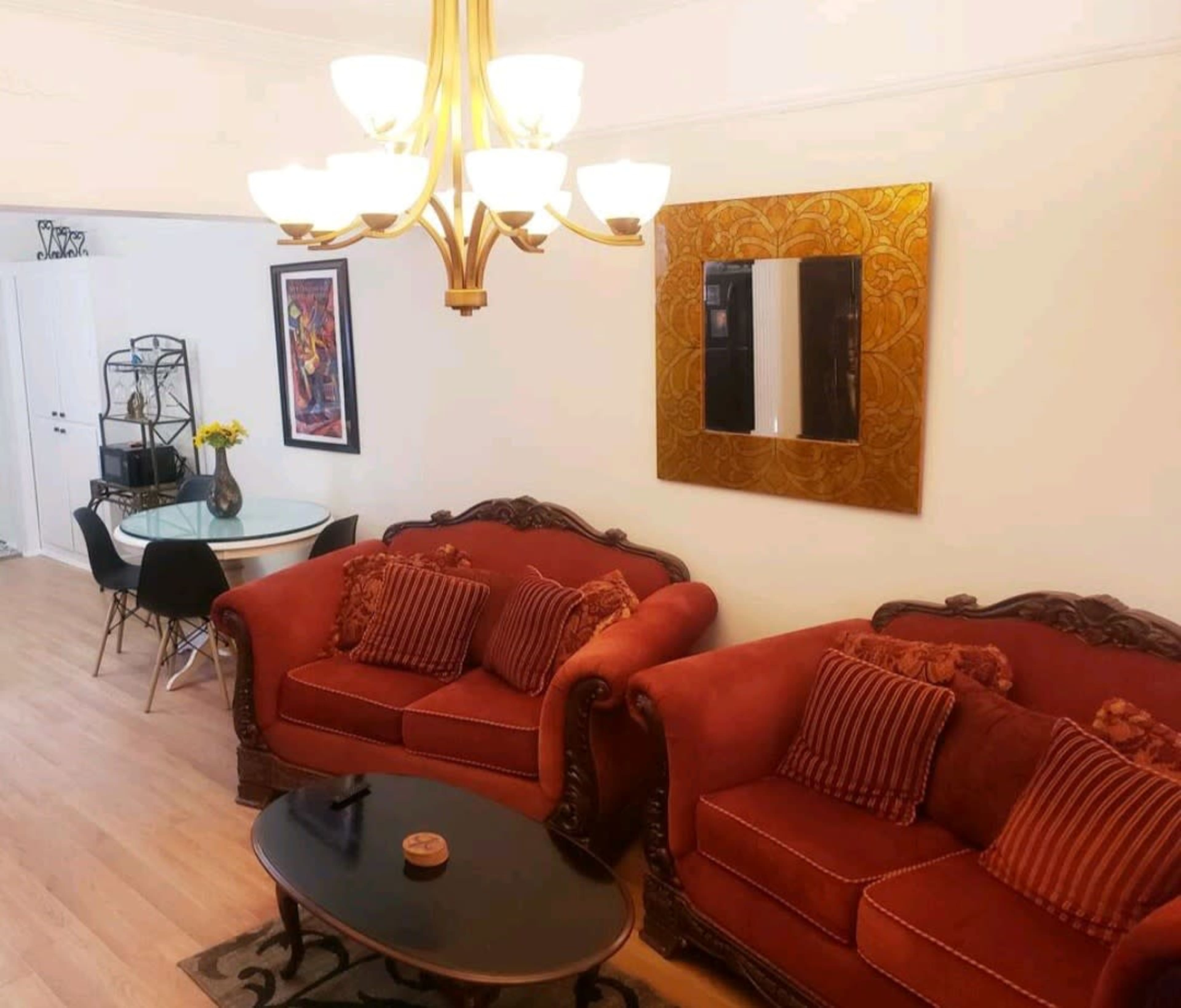 The living area features two red sofas with cushions, a round coffee table, and a chandelier, with a dining area and decorative mirror visible in the background.
