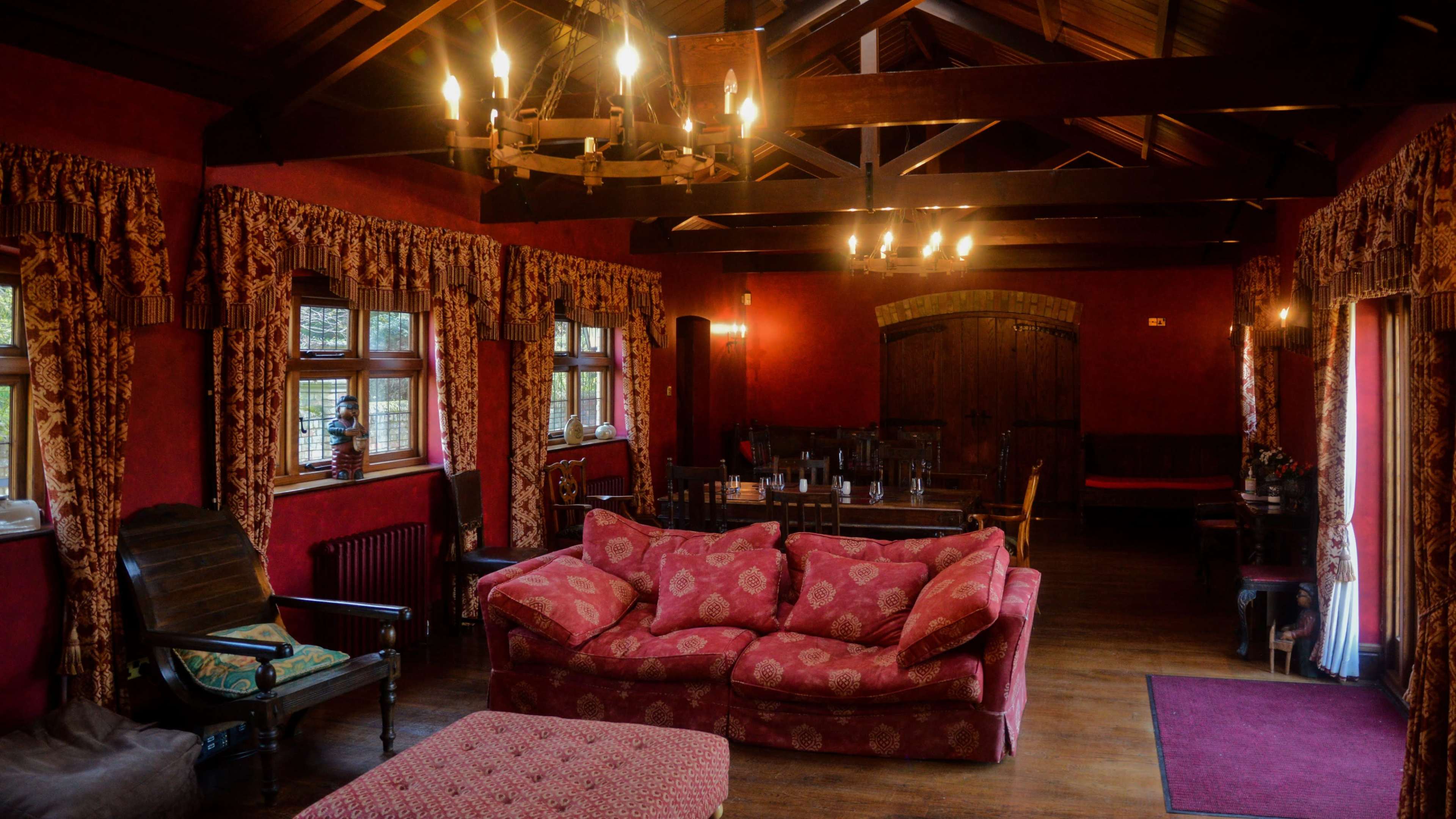The image shows a cozy room with wooden beams, red walls, a large upholstered sofa, and multiple windows that allow light to enter.