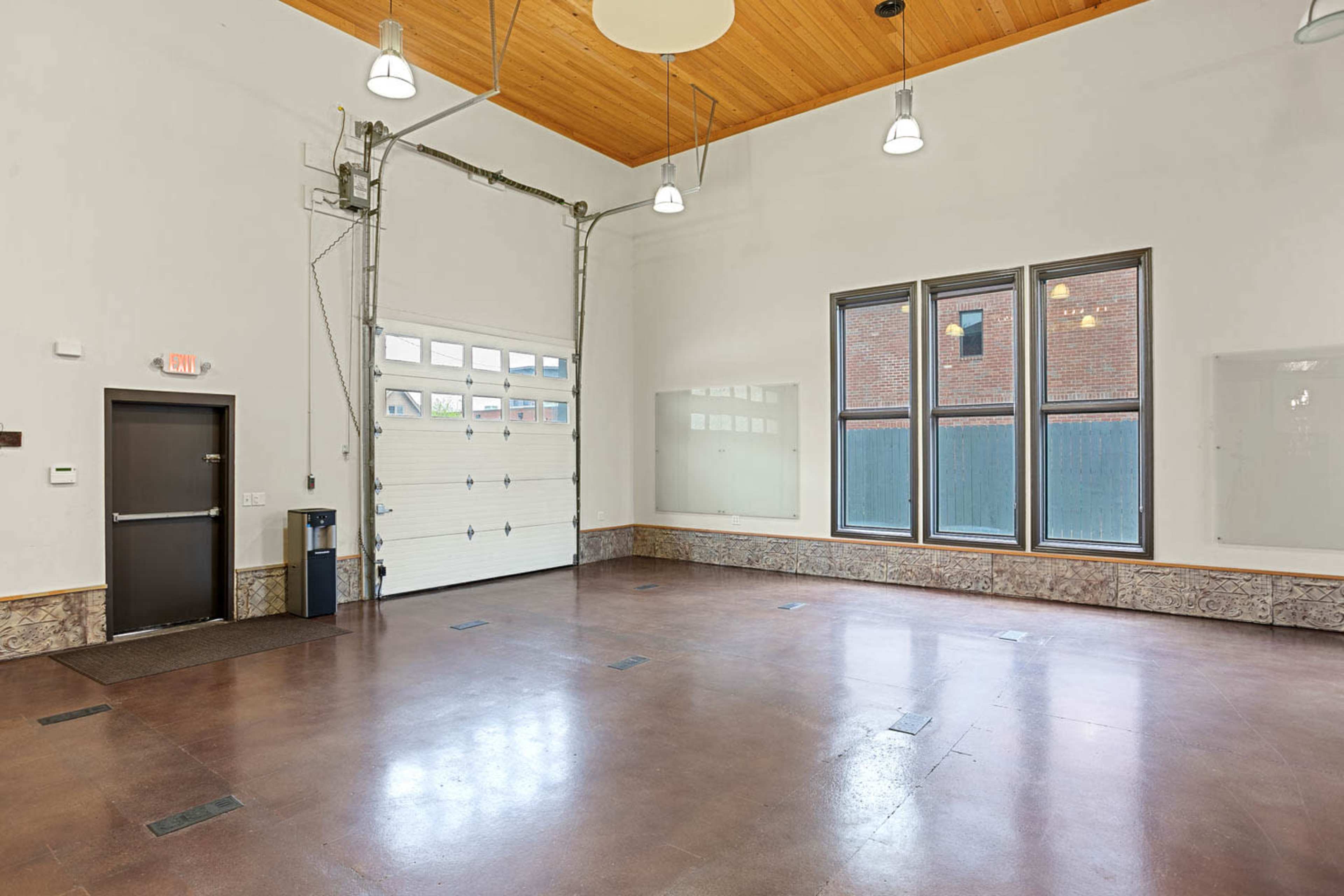 The image shows a spacious, empty room with large windows and a garage door, featuring a wood-paneled ceiling and concrete floors.