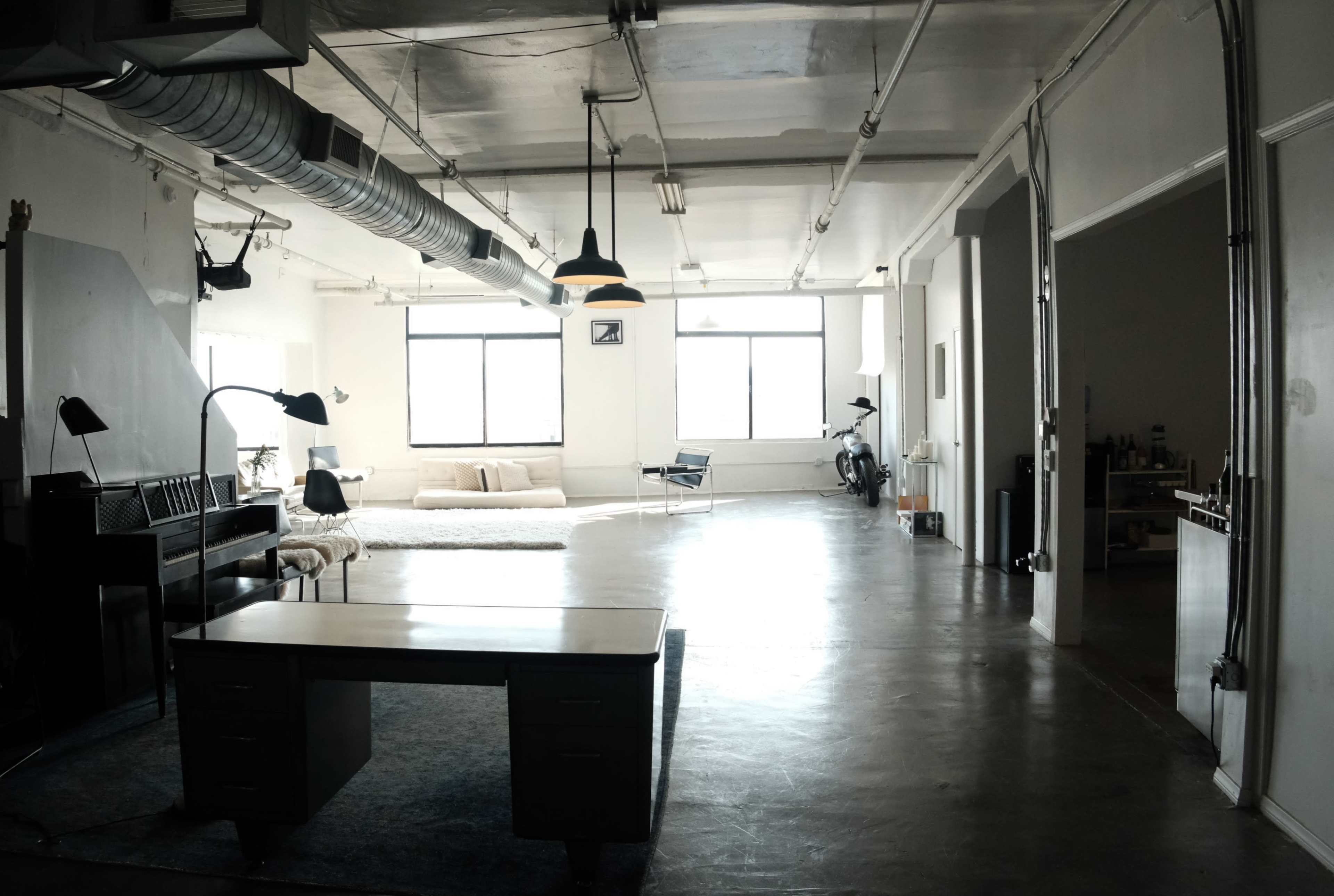 The image shows a spacious, minimally furnished loft with large windows, a desk, a seating area, and a motorcycle.