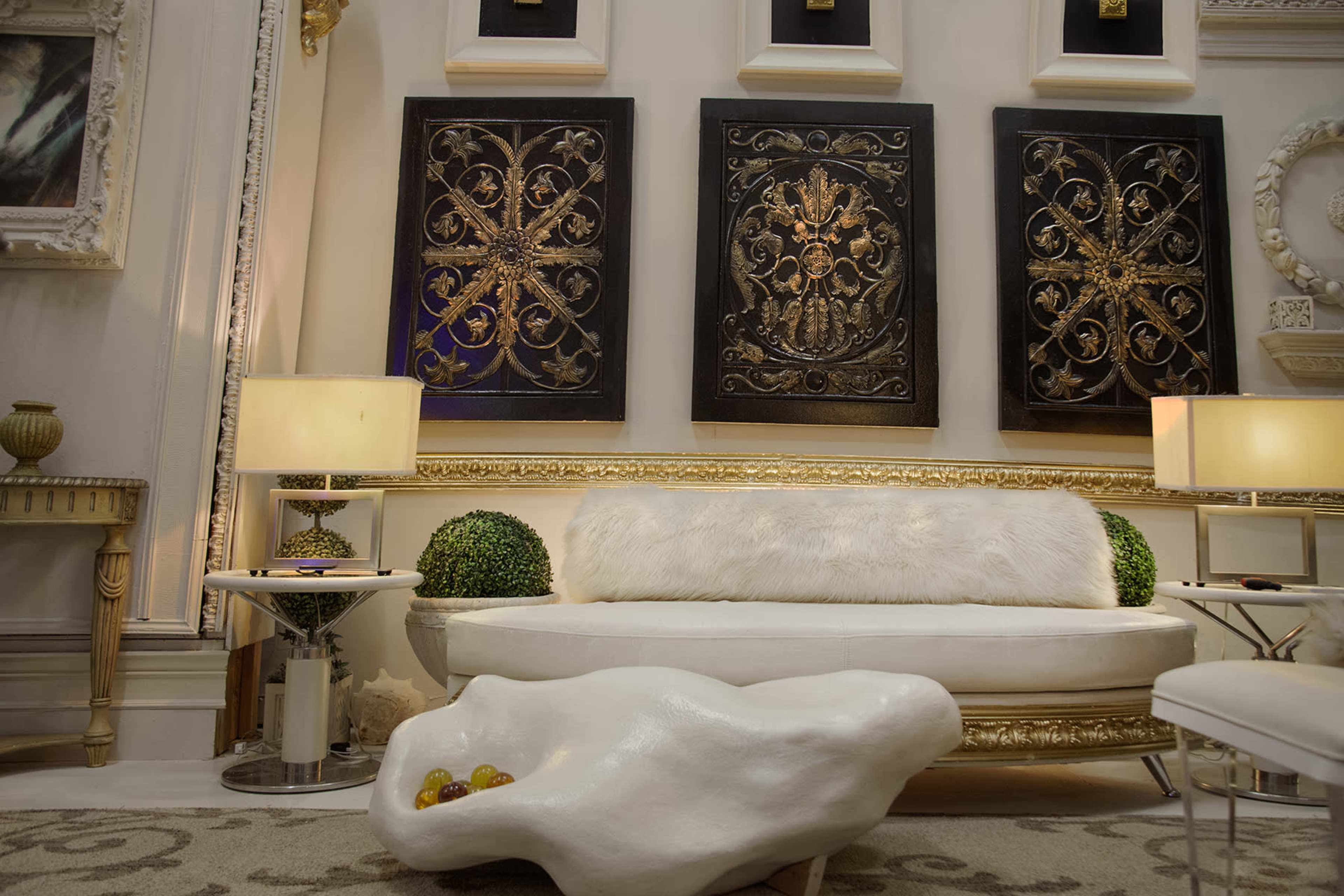 The image shows a stylish living space with a white sofa, decorative wall art with gold patterns, two lamps, and a unique sculptural coffee table.