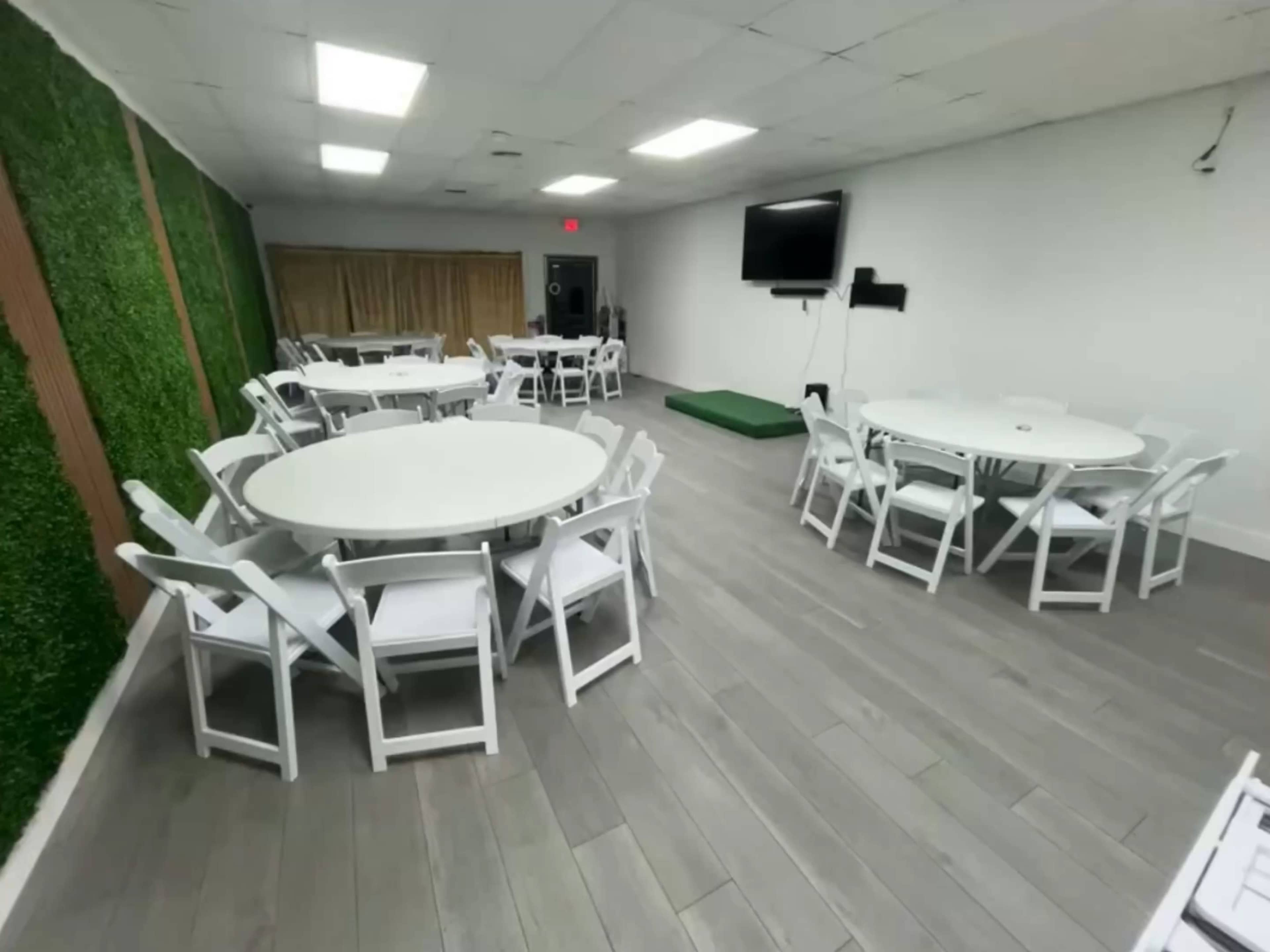 The image shows a spacious indoor room with white circular tables and chairs arranged neatly, surrounded by green wall decor and featuring a television mounted on a wall.