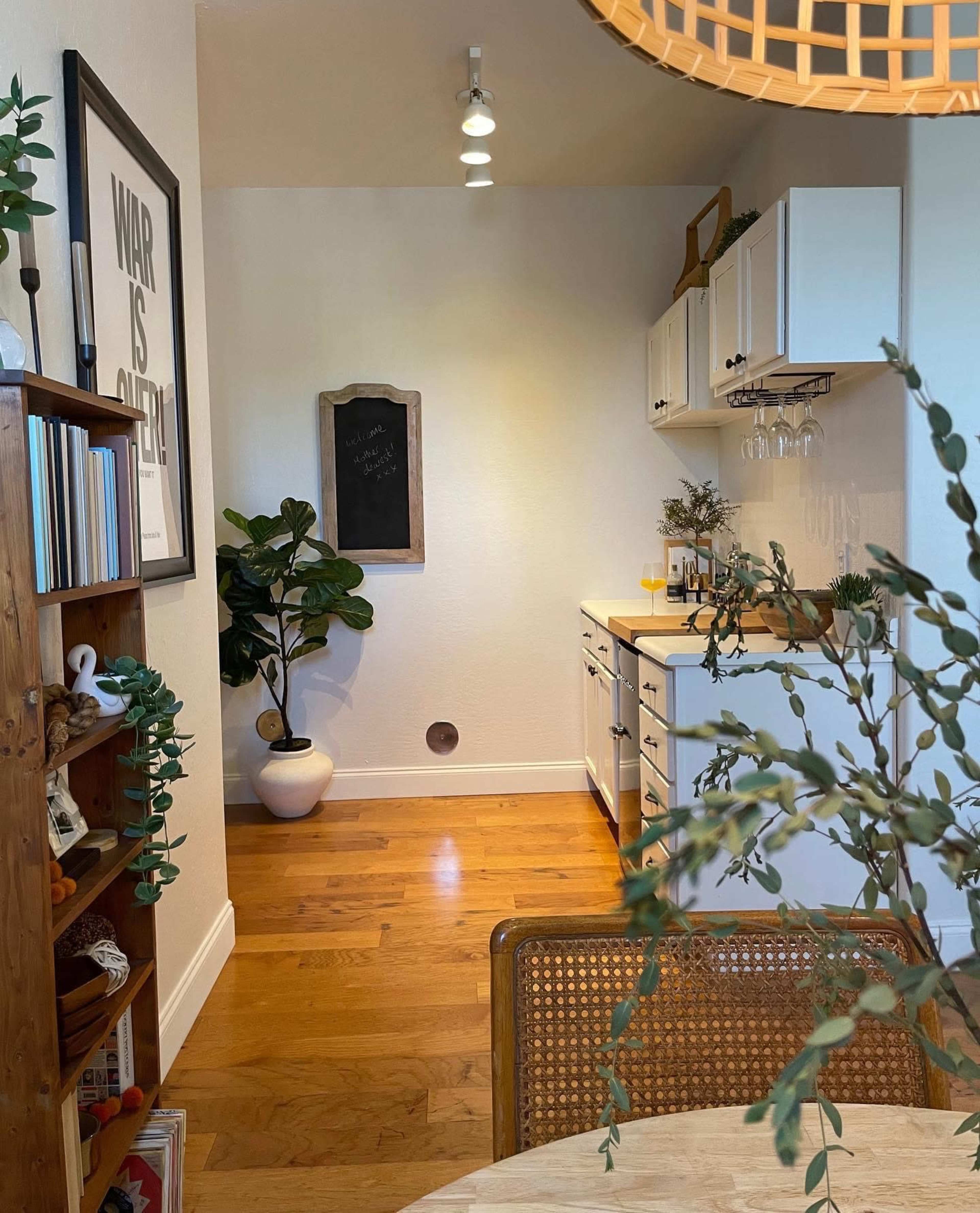 The image shows a modern kitchen area with white cabinetry, a chalkboard on the wall, and a potted plant in a bright, open space.