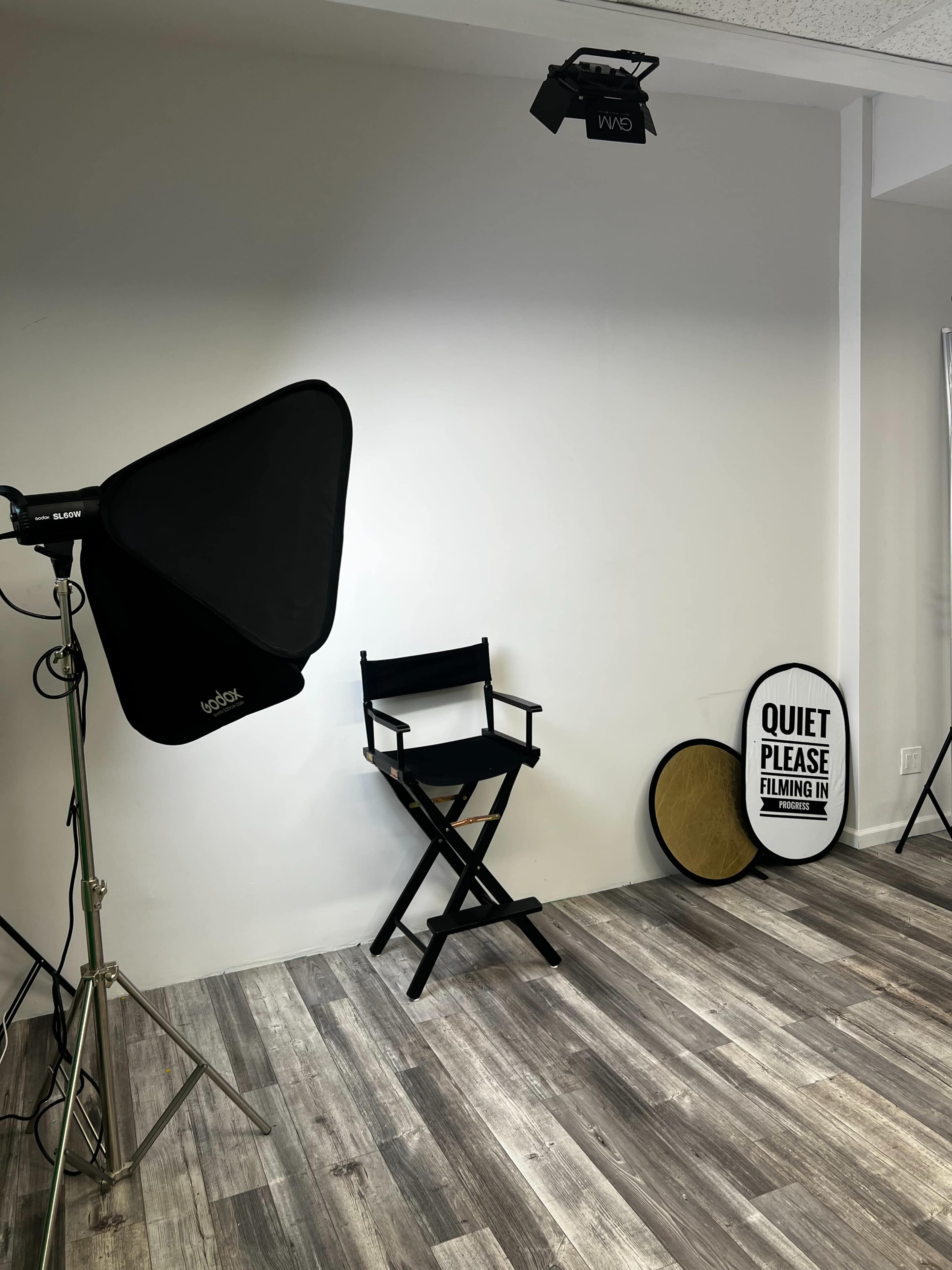 A film studio setup features a black director's chair beside softbox lighting, a sign that reads "QUIET PLEASE FILMING IN," and a circular reflector against a plain wall.