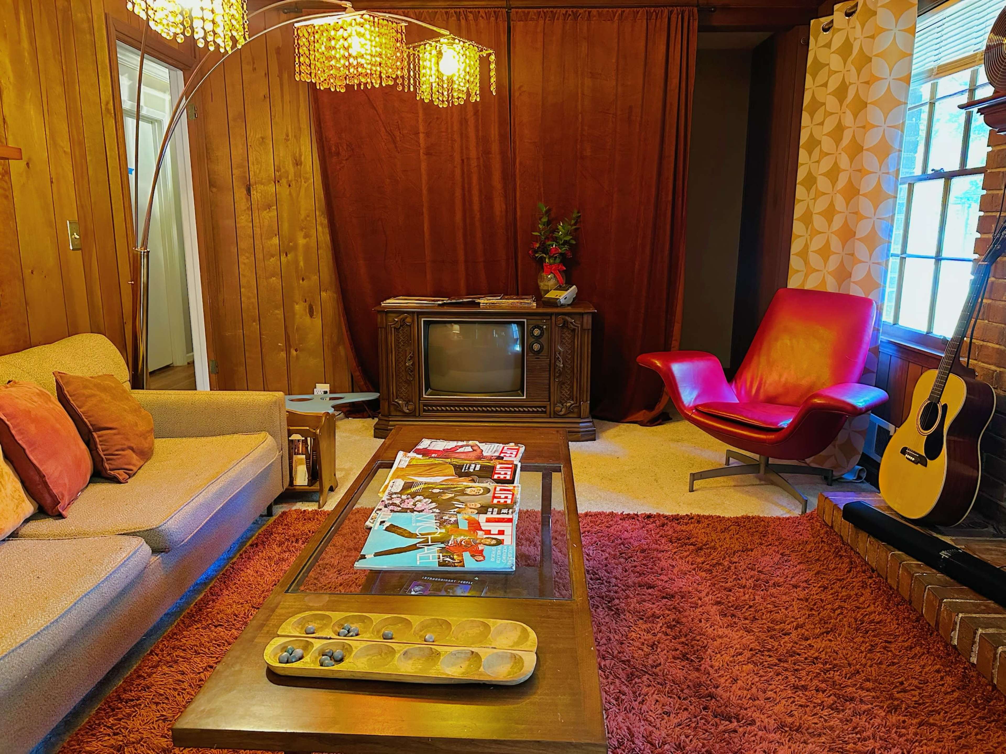 The image depicts a cozy living room featuring a retro television, a red lounge chair, a wooden coffee table with decorative items, and a warm-toned rug under soft lighting.