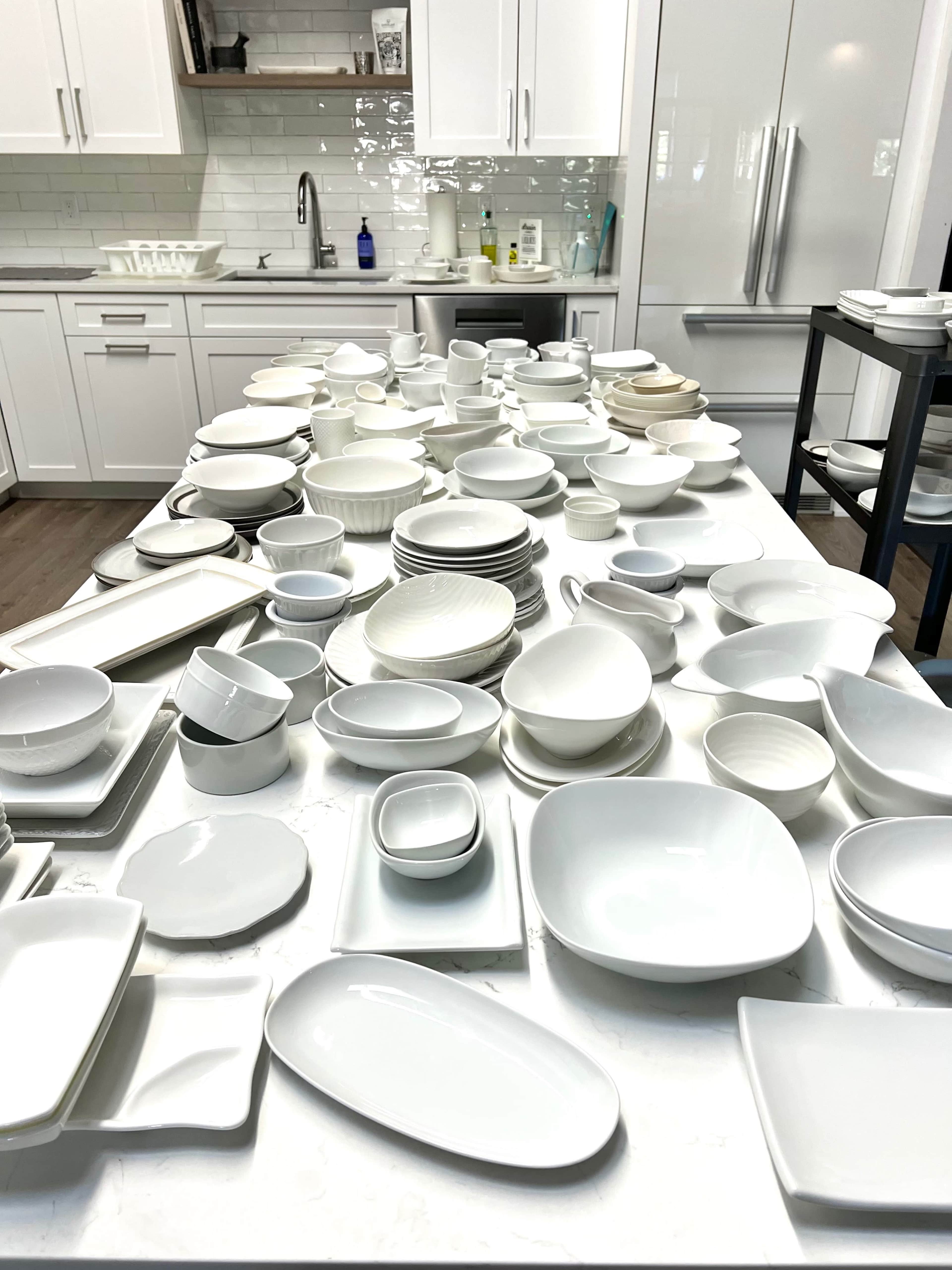 A large assortment of white dishware and kitchenware is spread out on a table in a modern kitchen.