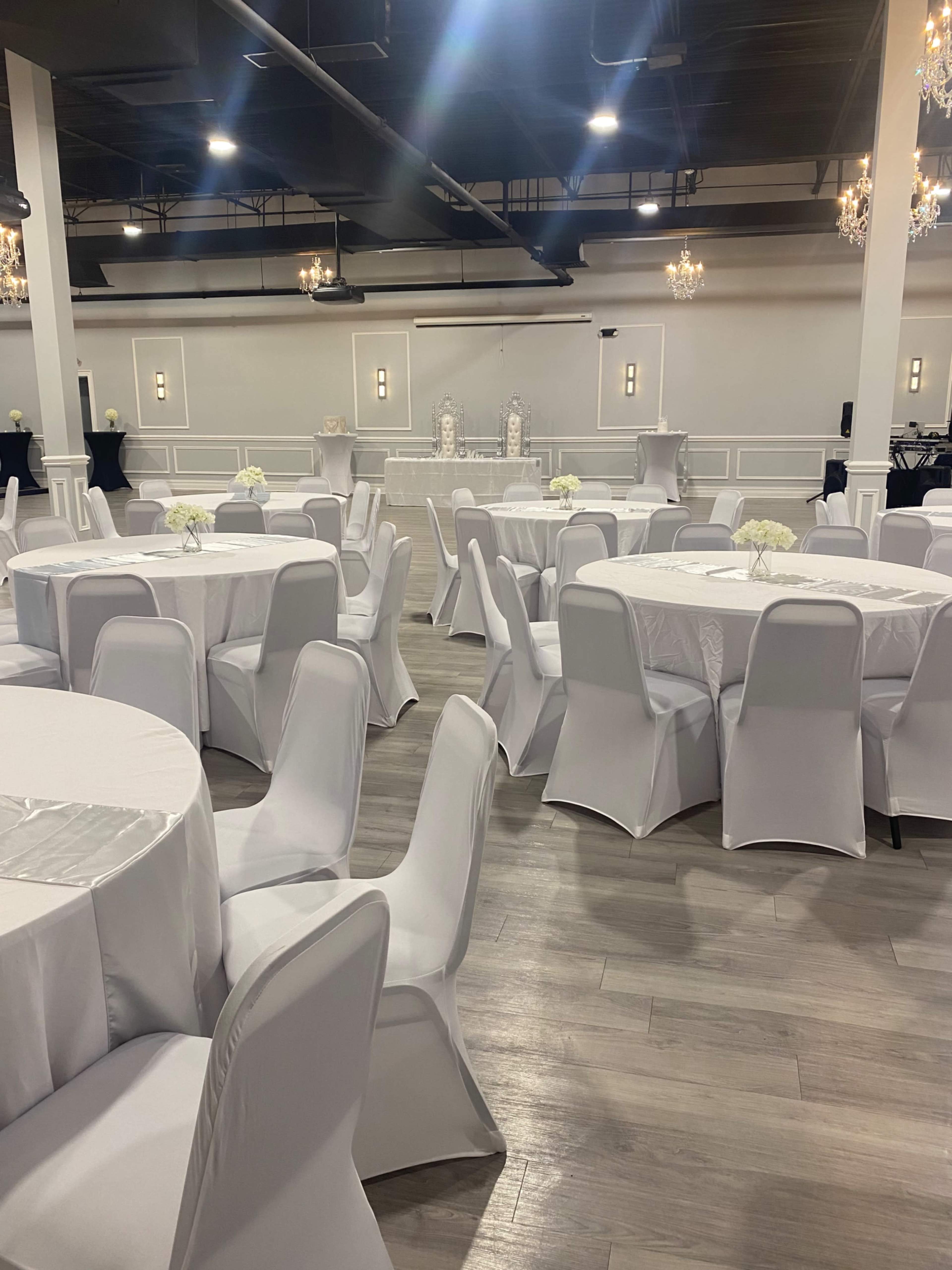 A large event space with round tables covered in white tablecloths and white chairs, arranged in a setup for an event, with chandeliers hanging above.