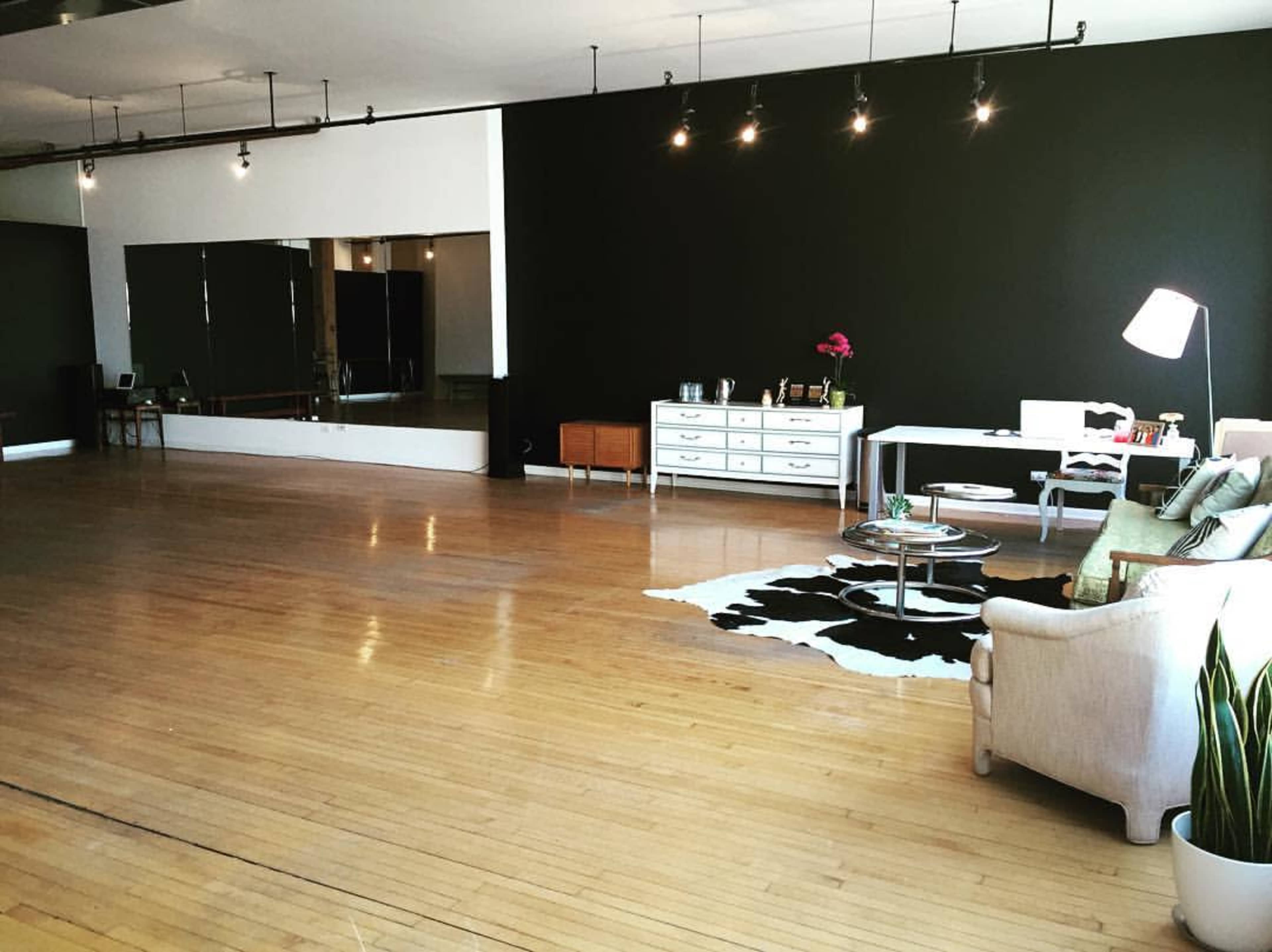 The image depicts a spacious studio with dark green walls, wooden flooring, a seating area, a large mirror, and a desk against one wall.