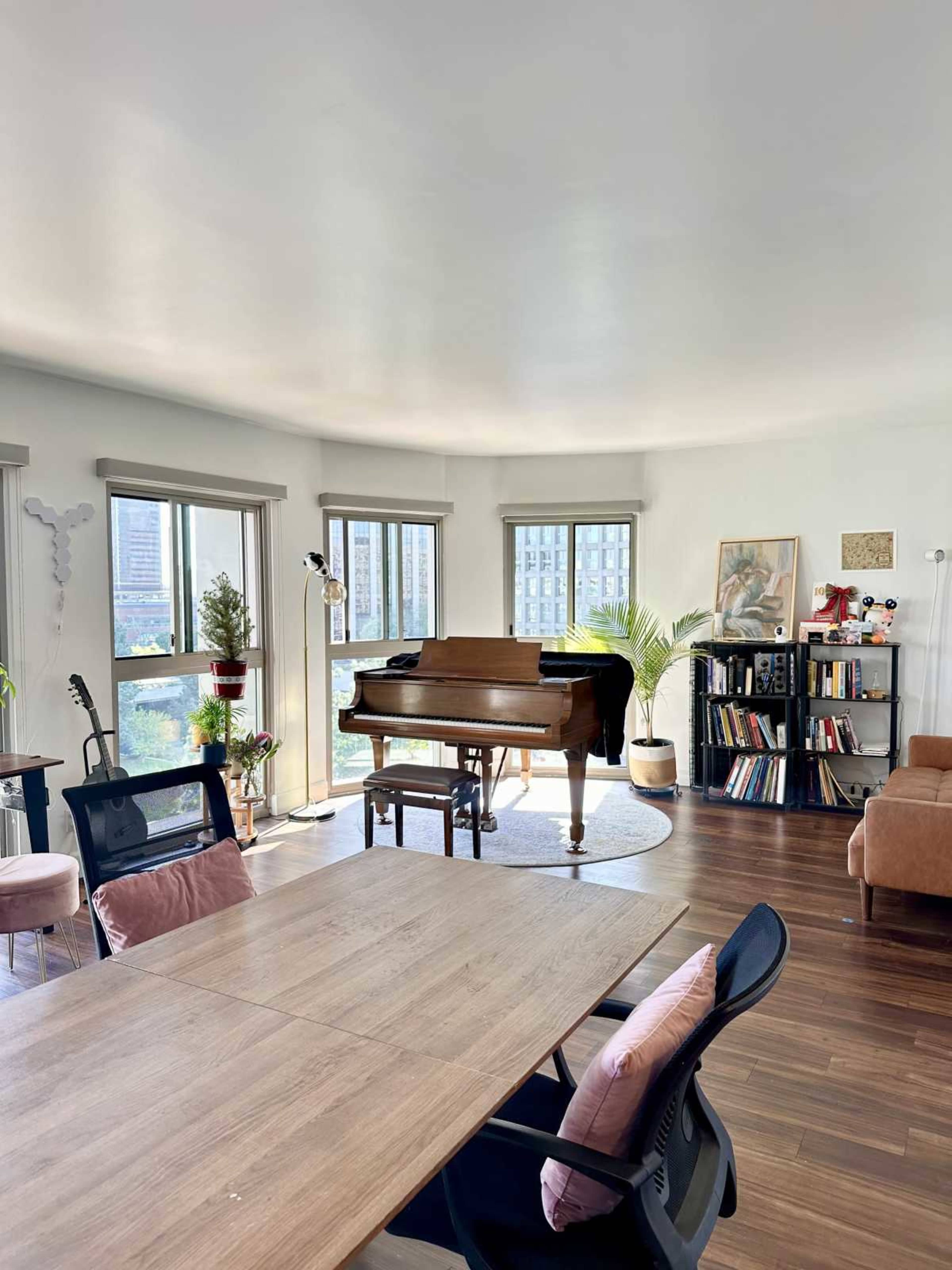 The image shows a bright, modern living room featuring a grand piano, a dining table, plants, and a bookshelf.