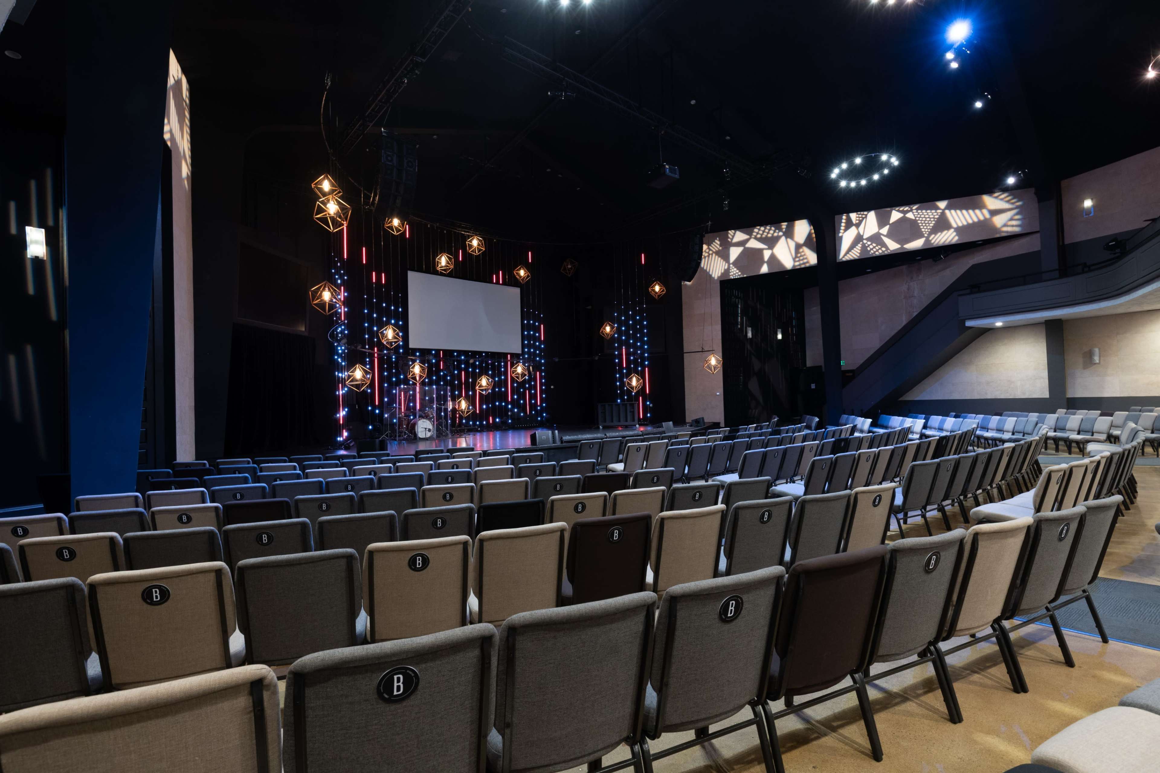 The image shows an auditorium with rows of seats facing a stage adorned with lights and a large screen.