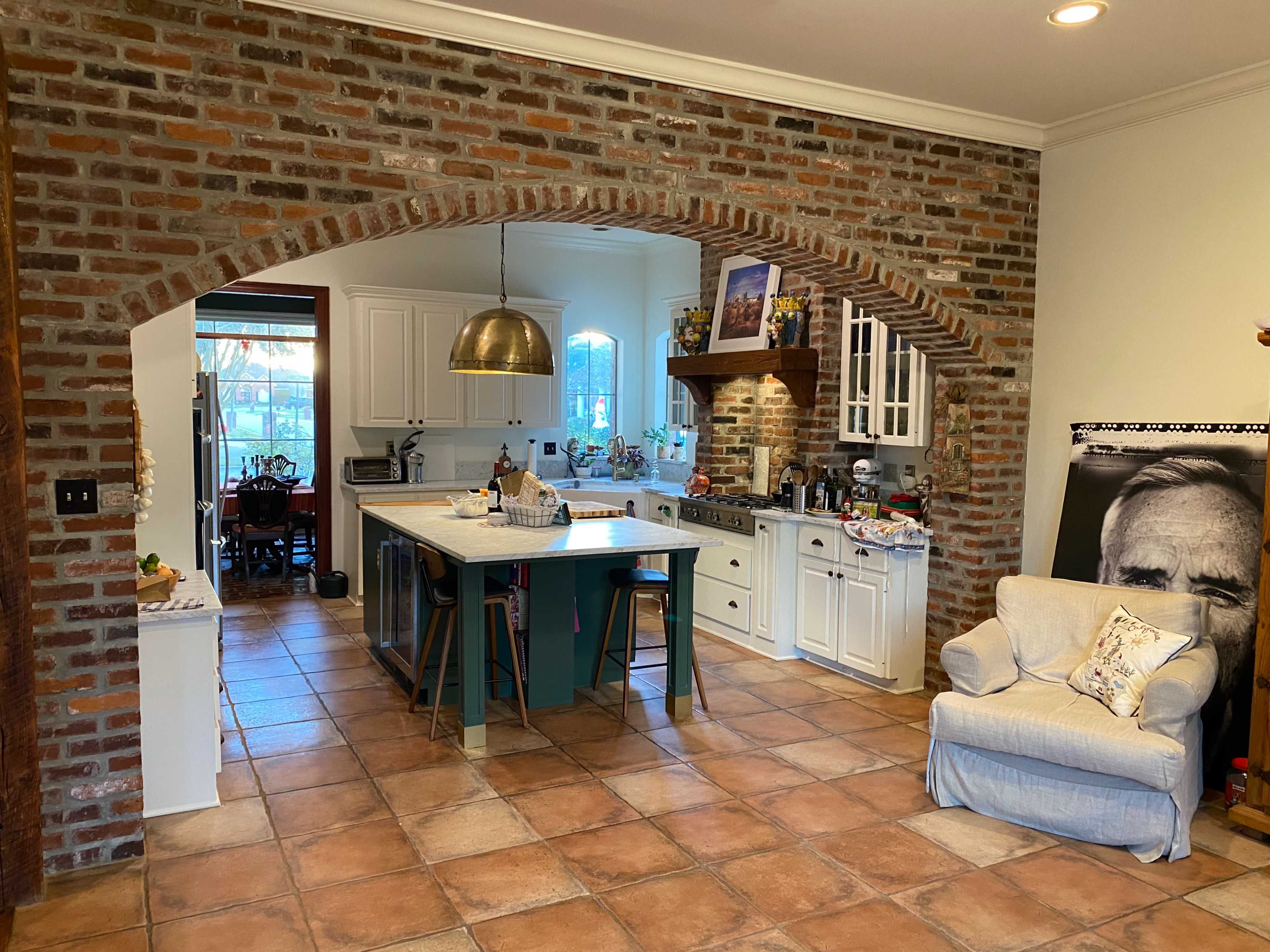 A spacious kitchen with a brick archway, white cabinetry, an island with a turquoise base, and a cozy seating area featuring a light blue armchair.