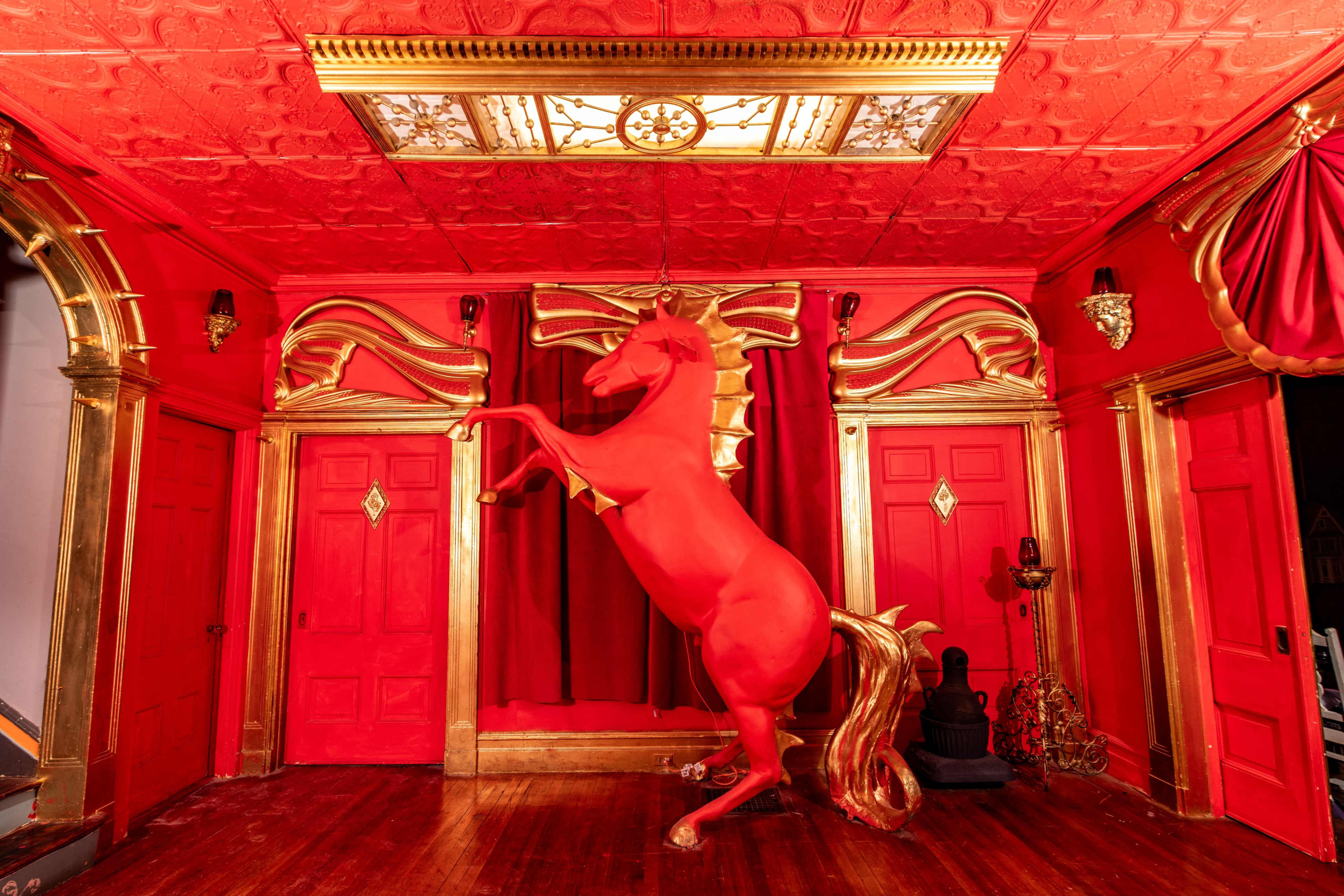 The scene features a vividly red room adorned with ornate gold decorations and a life-sized statue of a rearing horse at the center.