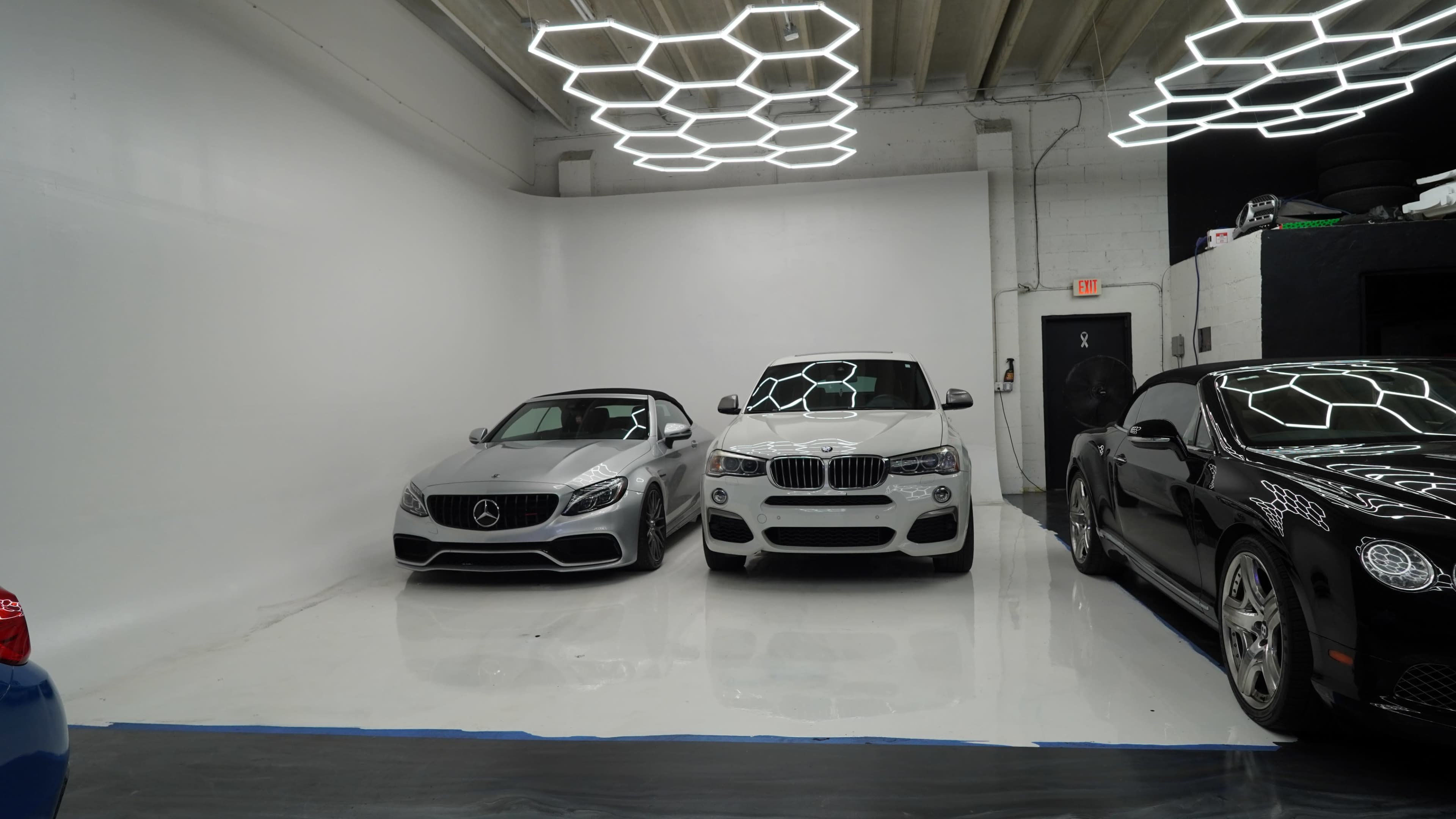 The image shows a modern garage with three luxury cars: a silver Mercedes convertible, a white BMW SUV, and a black Bentley.