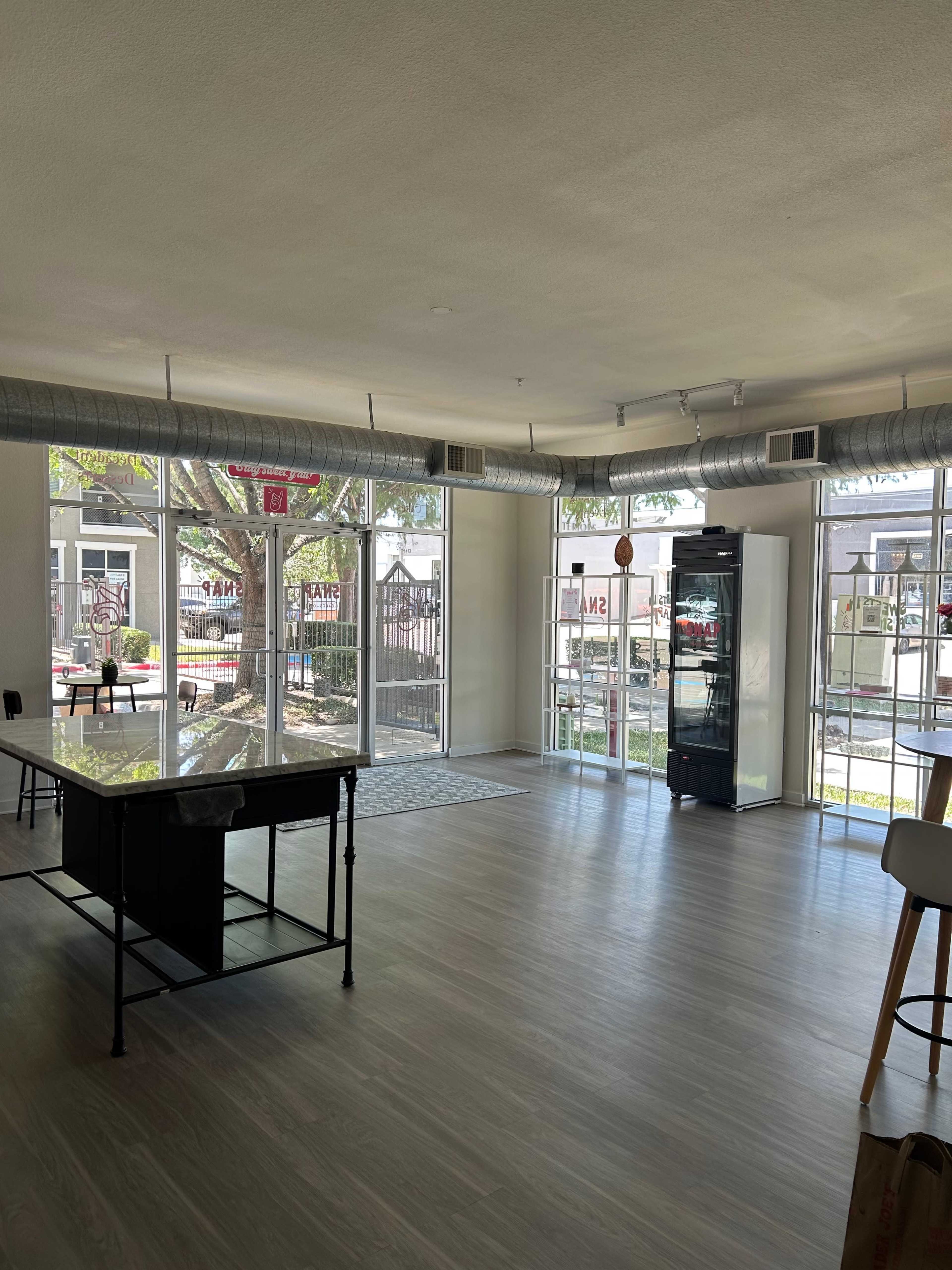 The image shows an empty modern indoor space with large windows, a counter with stools, and a refrigerator against one wall.