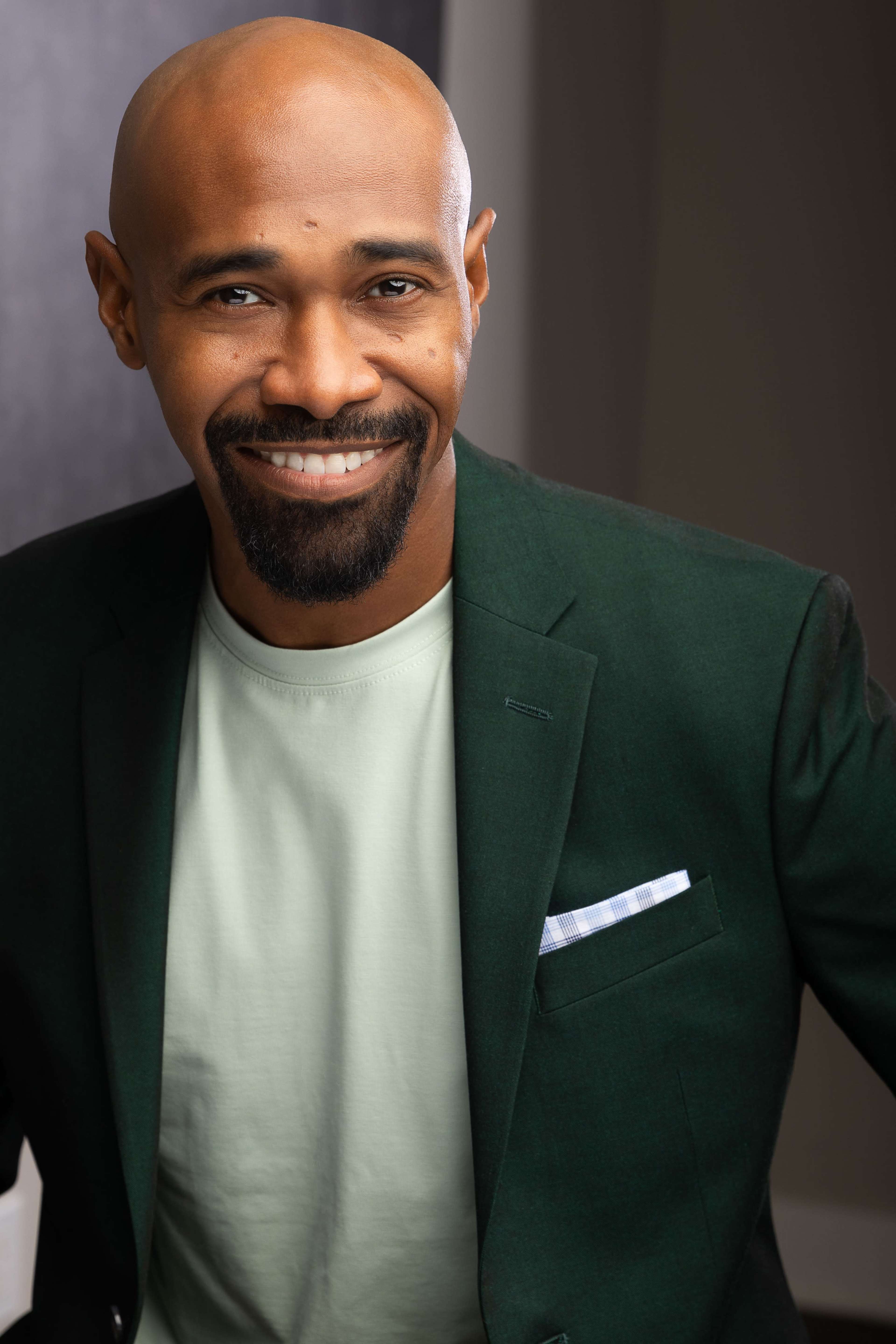 A man with a shaved head and a well-groomed beard is smiling while wearing a dark green blazer over a light green shirt.