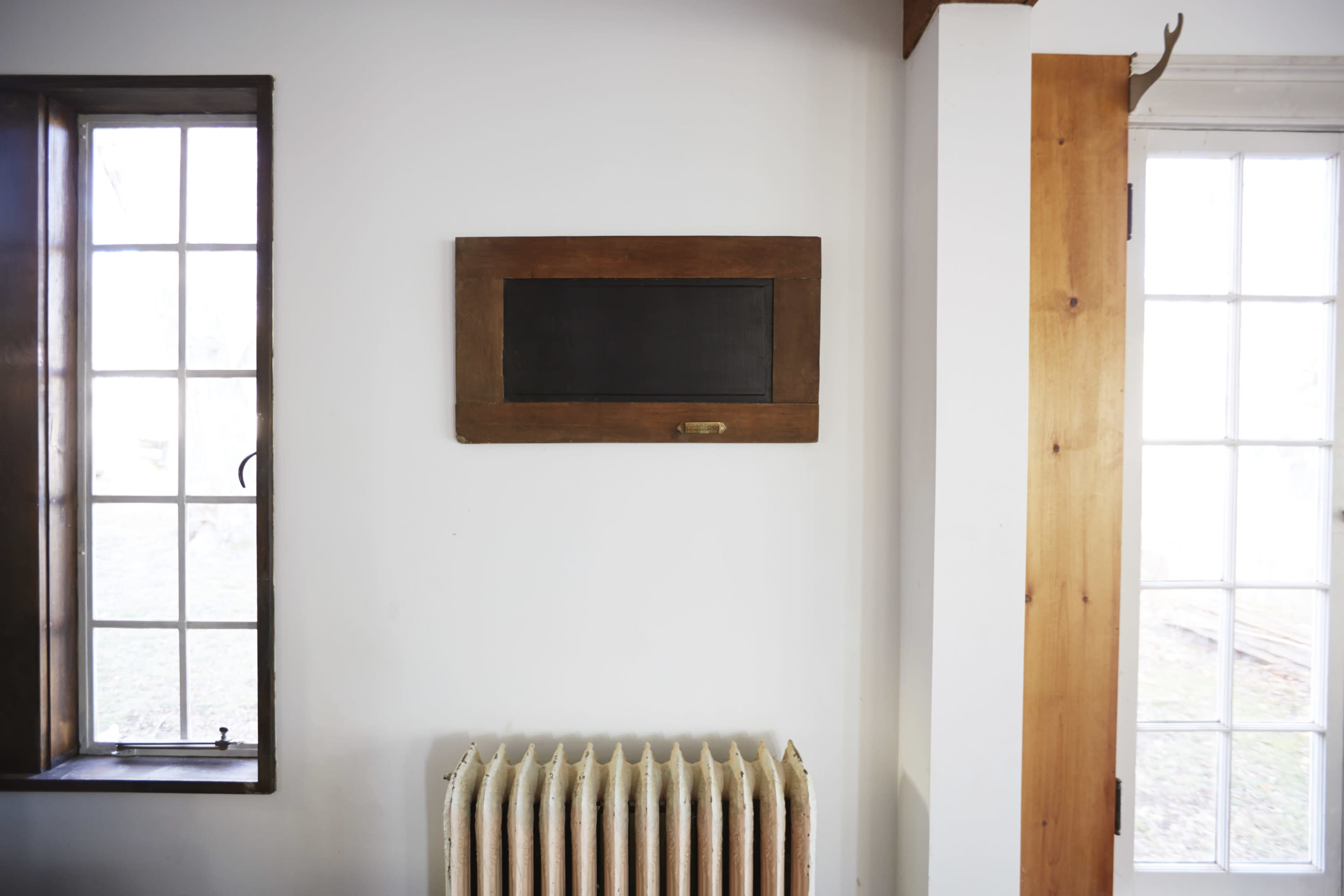 A simple interior space featuring a chalkboard mounted on a white wall, a vintage radiator below, and windows on either side.