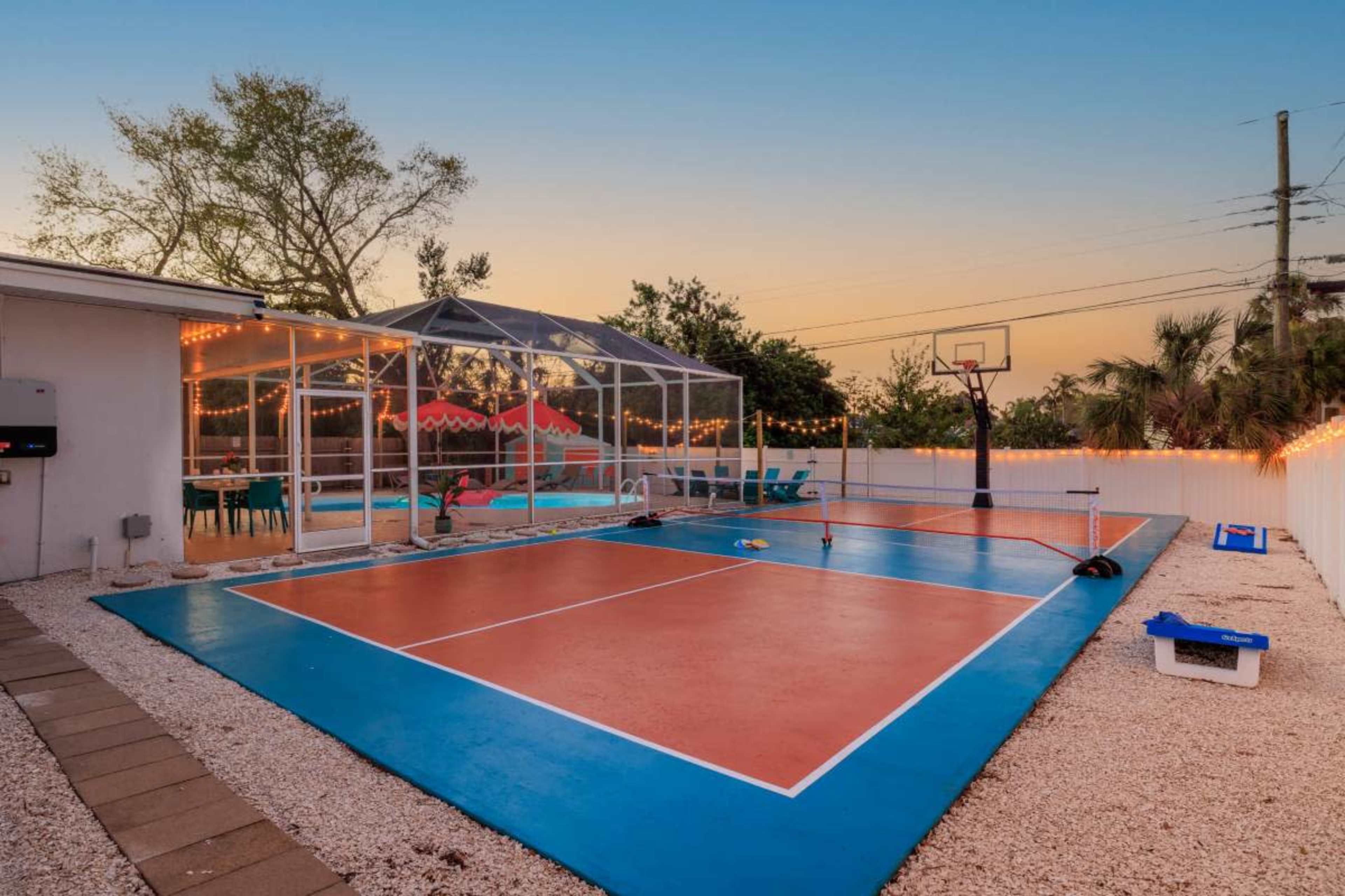 The image shows a fenced outdoor sports court with a basketball hoop and a net area, surrounded by landscaping and a clear evening sky.