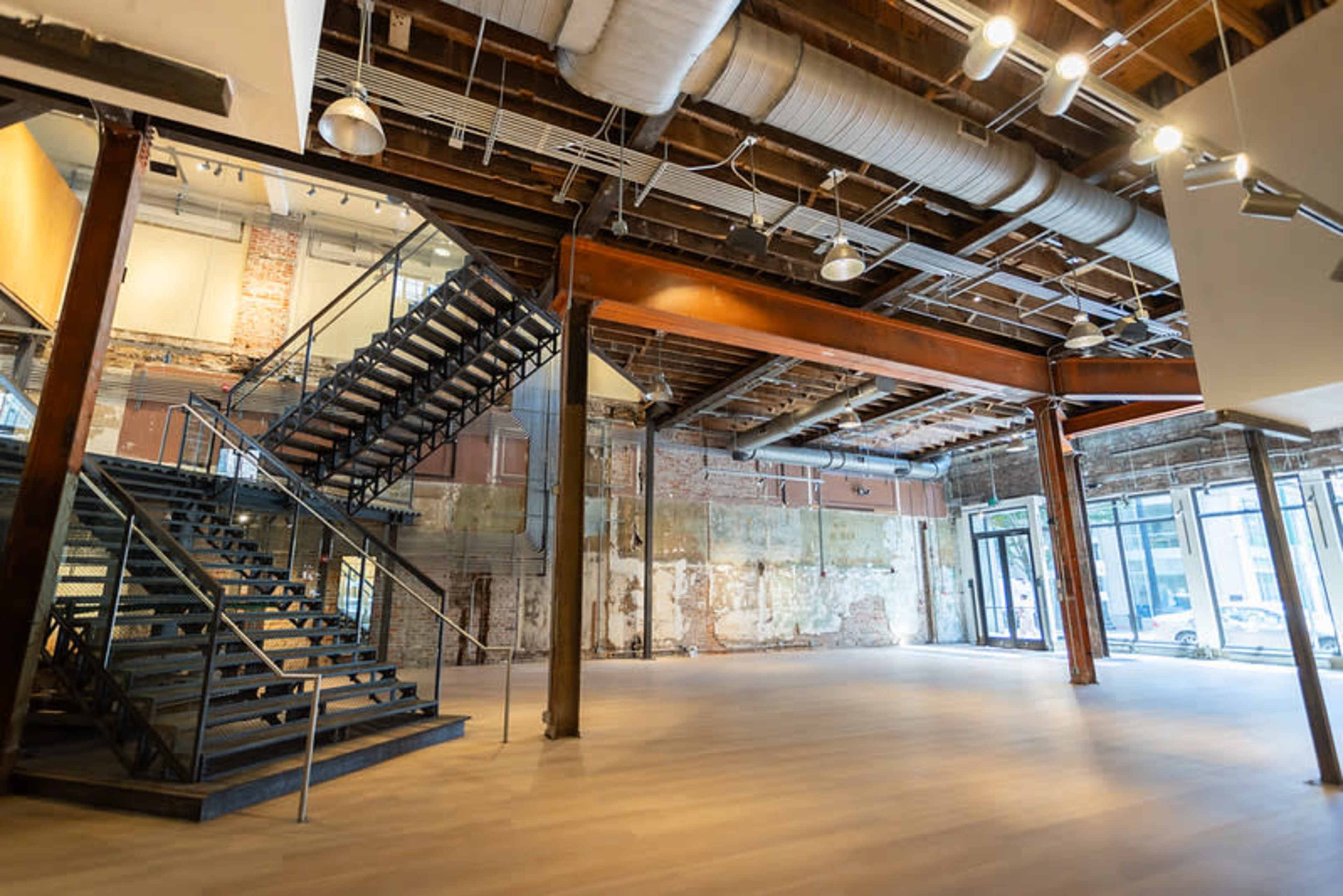 The image shows an interior view of a spacious, industrial-style room featuring exposed brick walls, a staircase with metal railings, and large windows.