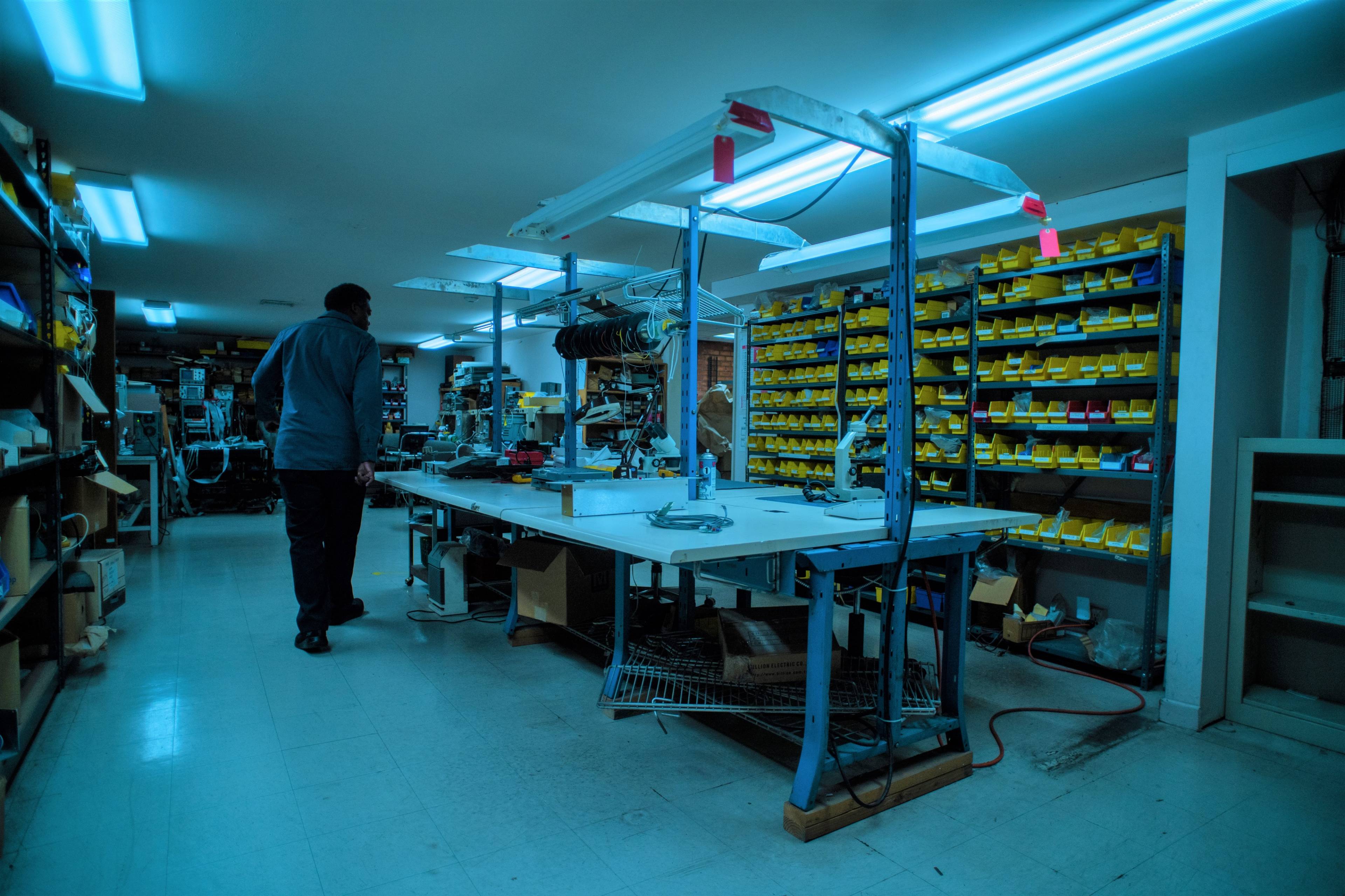A person walks through a dimly lit workshop filled with tools, equipment, and neatly organized yellow storage bins along the walls.