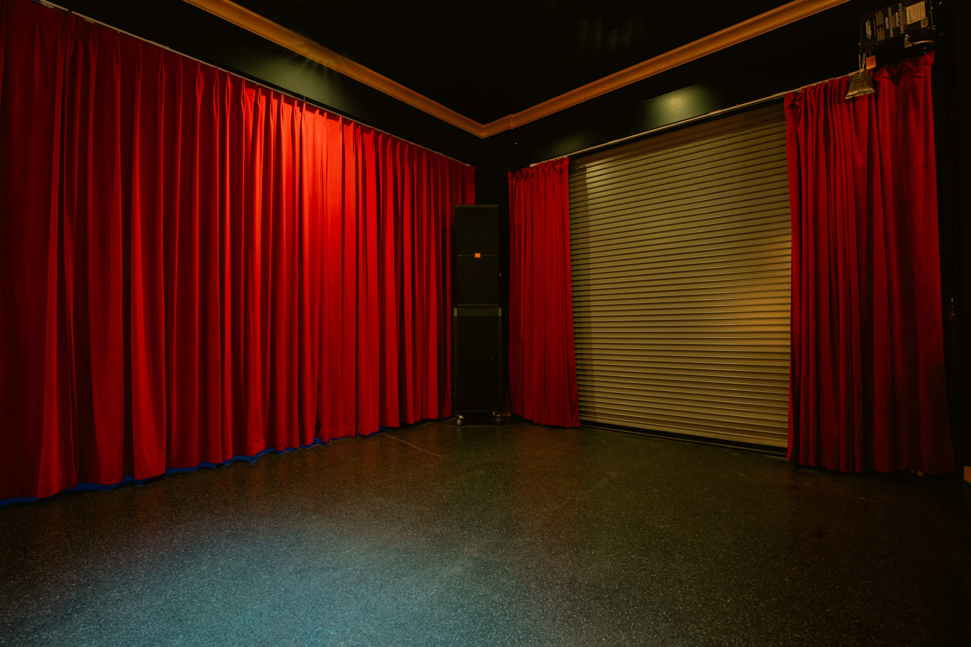 A dimly lit room with red curtains, a closed metal garage door, and a speaker system in the corner.