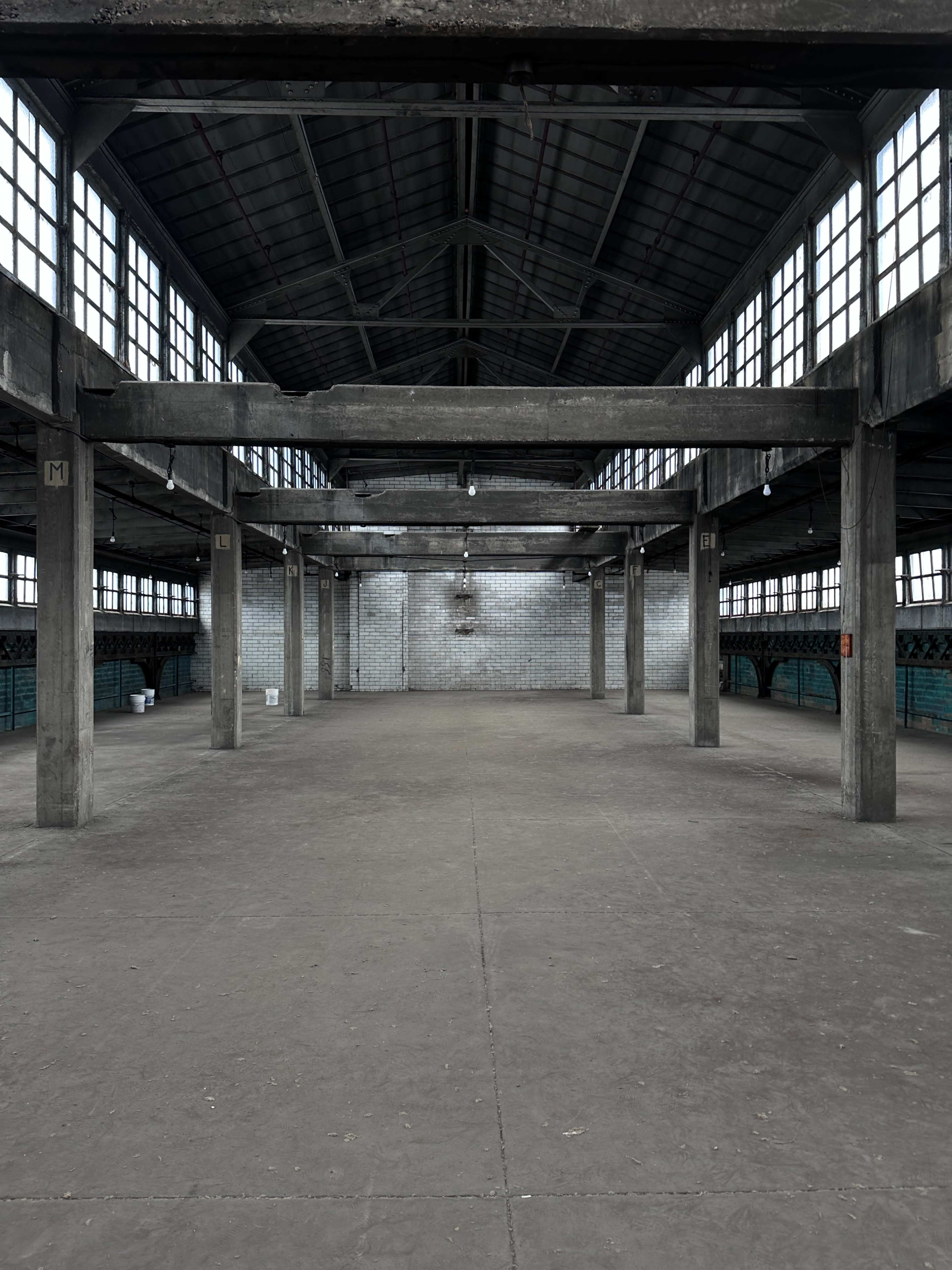An expansive, empty industrial warehouse features concrete floors and tall, windowed walls supported by large columns.