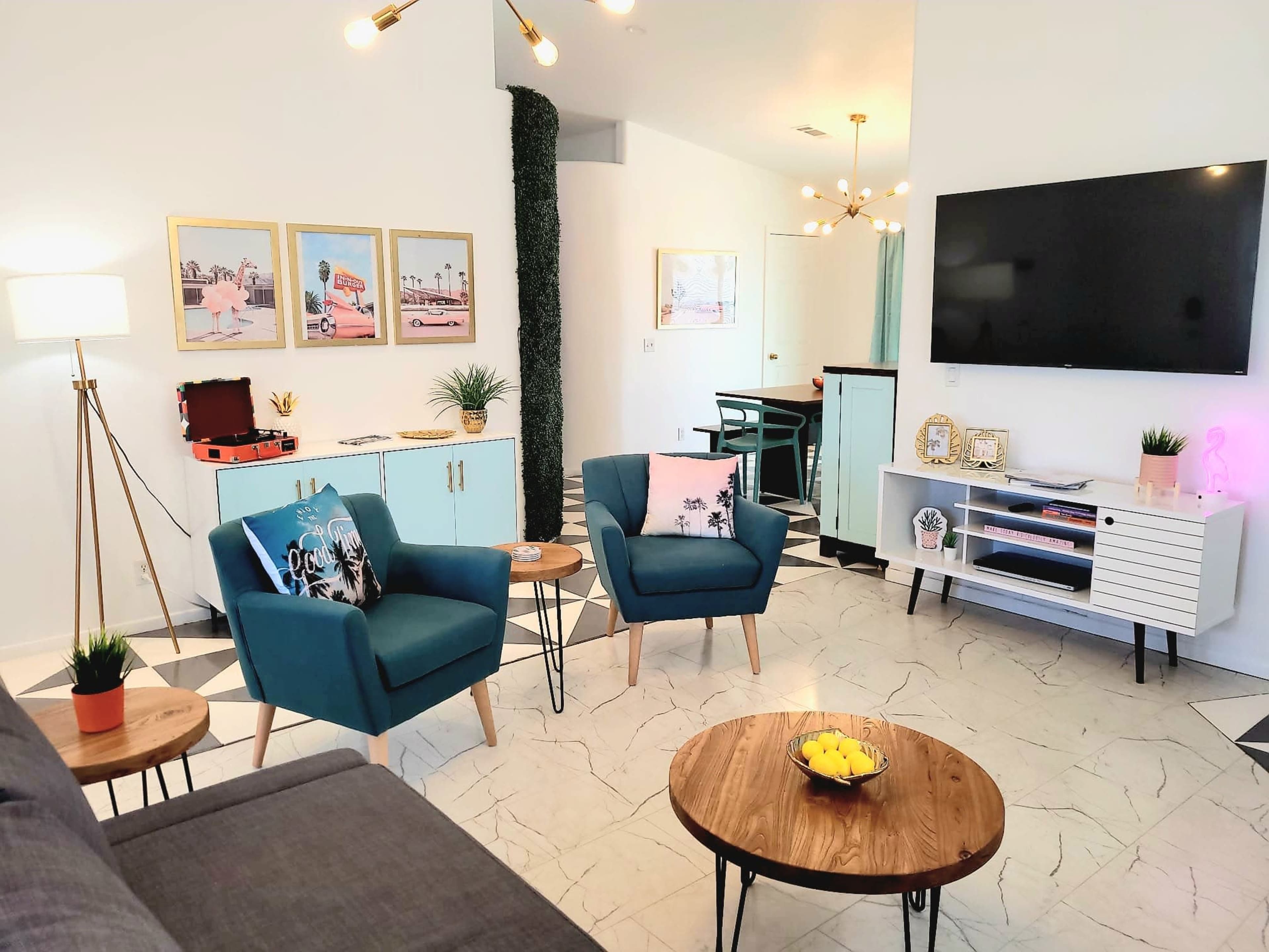 The image shows a modern living room with two teal chairs, a wooden coffee table, and a large television mounted on the wall, surrounded by decorative elements and art.