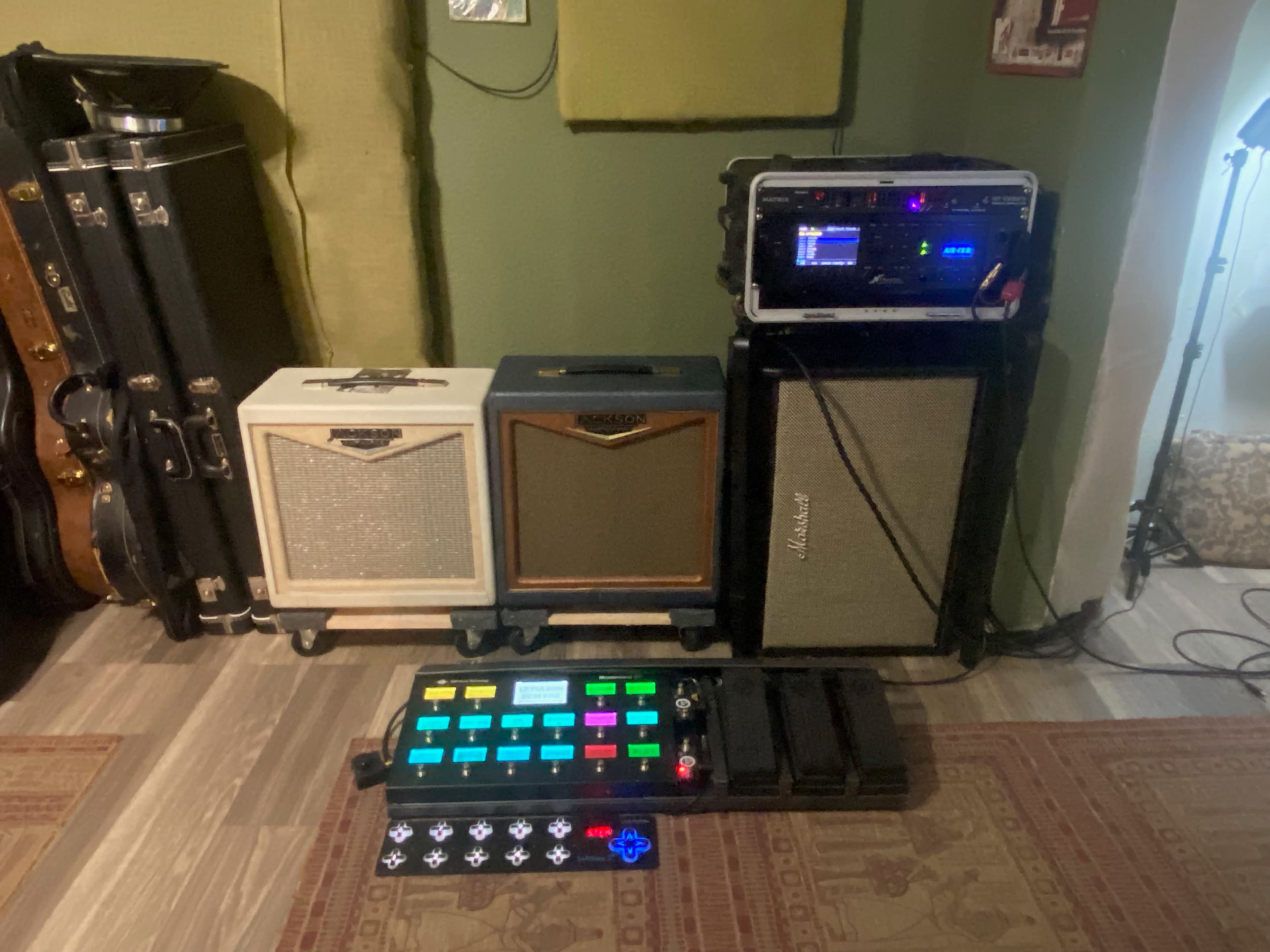 A setup featuring three amplifiers stacked on a wall in a music studio, with a multi-effects guitar pedalboard positioned on the floor in front.