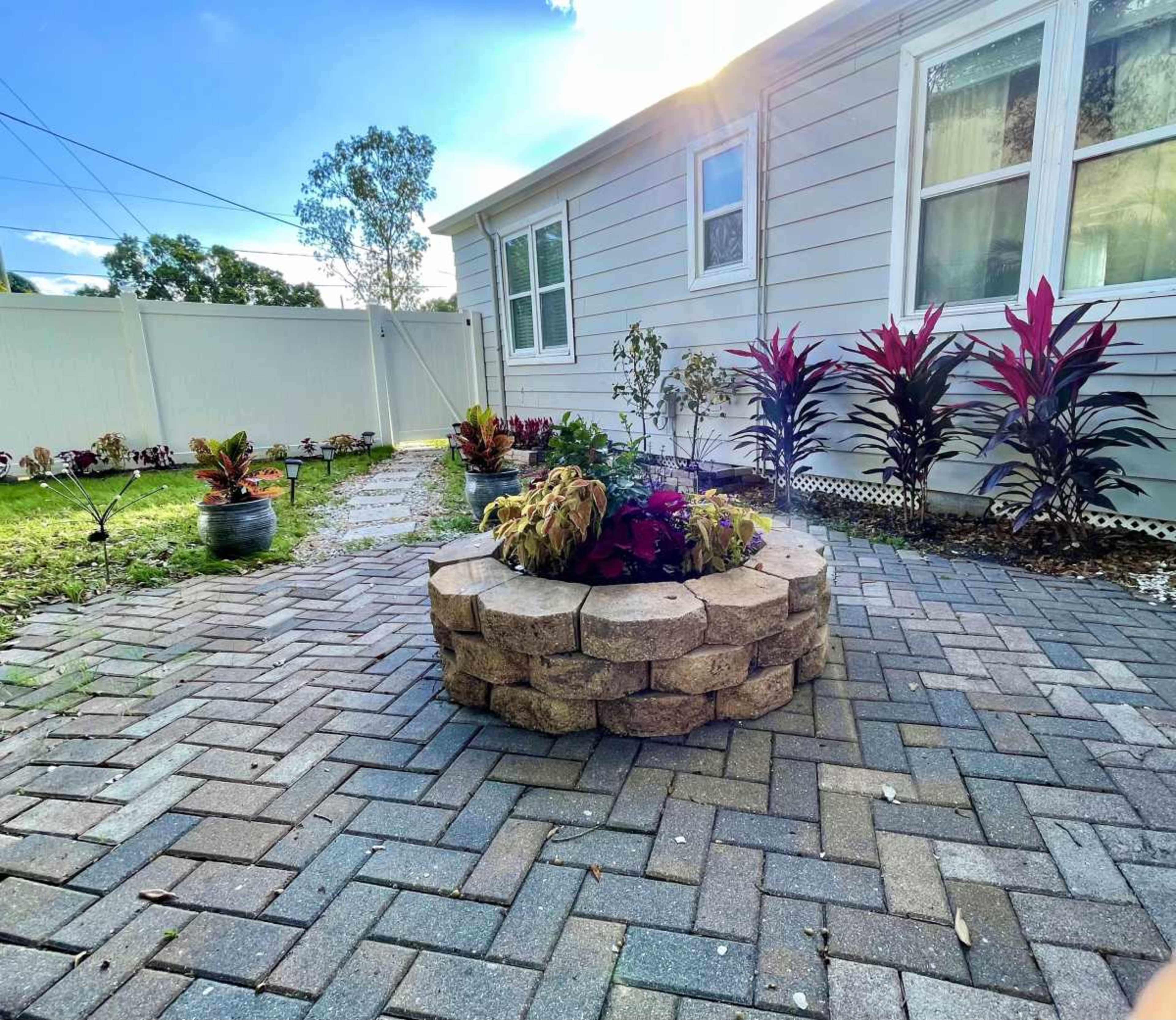 The image shows a patio area with a stone circular planter surrounded by pavers, colorful plants, and a house in the background.