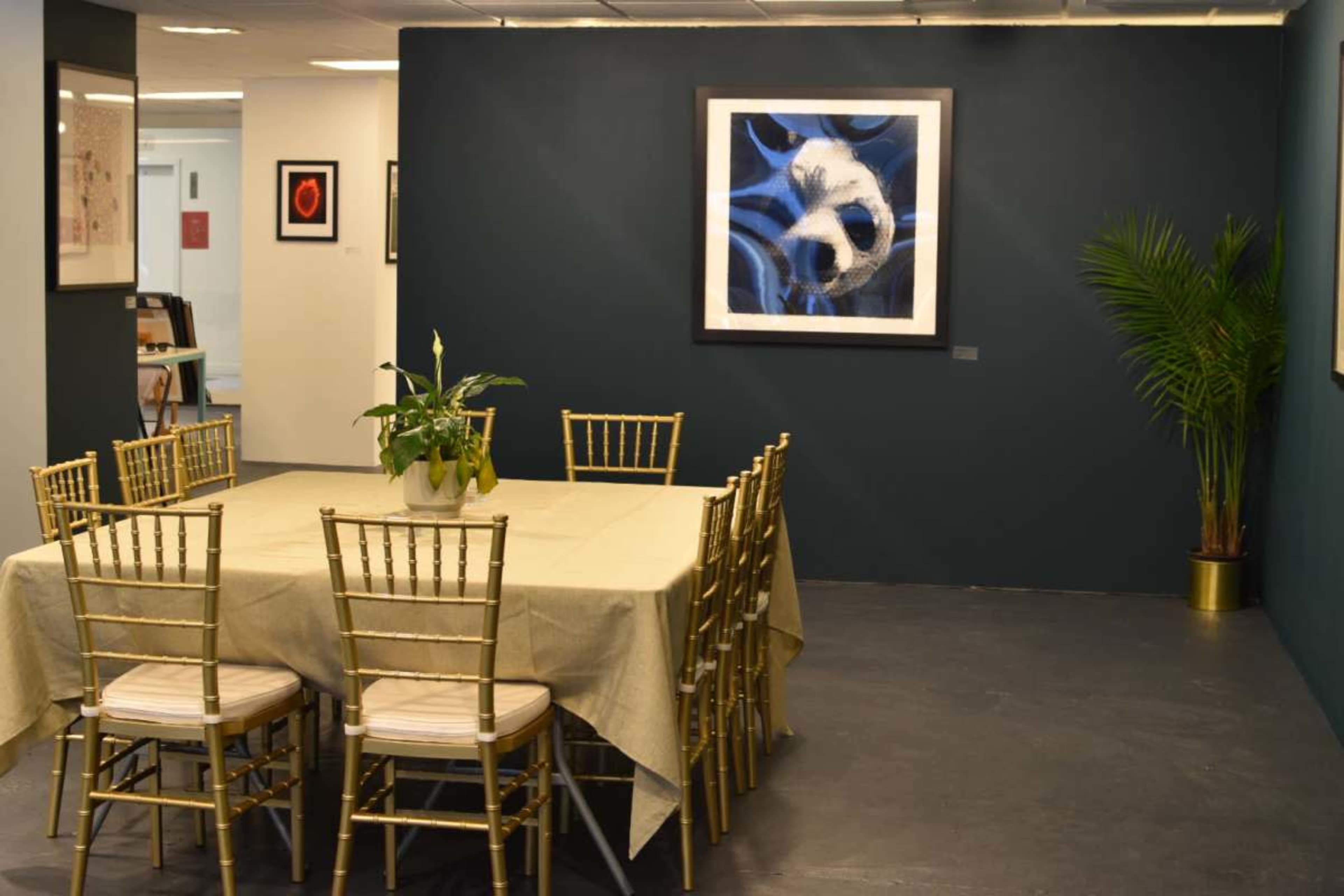 The image shows a well-lit room featuring a large rectangular table surrounded by gold-painted chairs, with a plant and a framed artwork on the dark green wall.