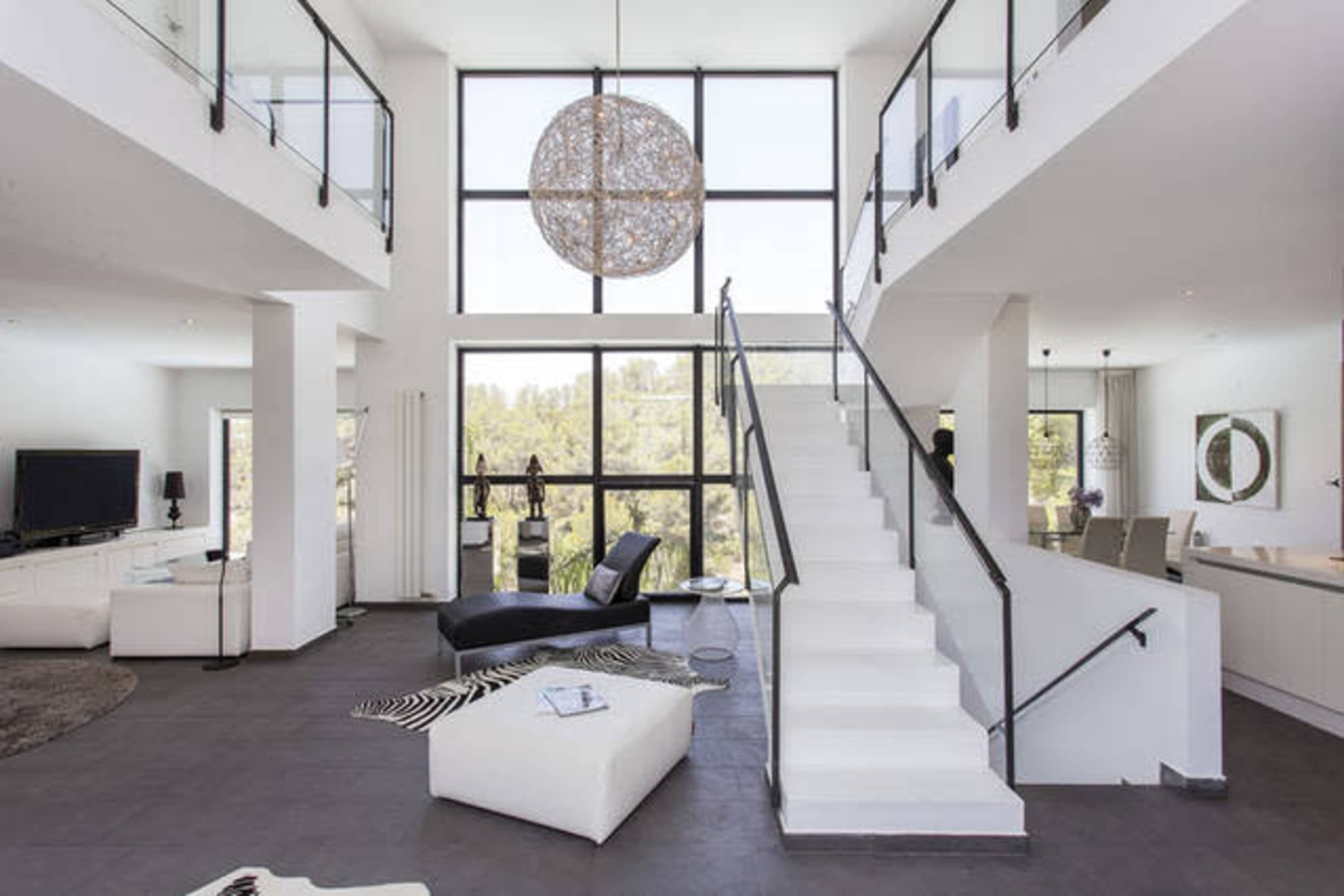 A modern, two-story living room with large windows, a staircase, and sleek furniture.