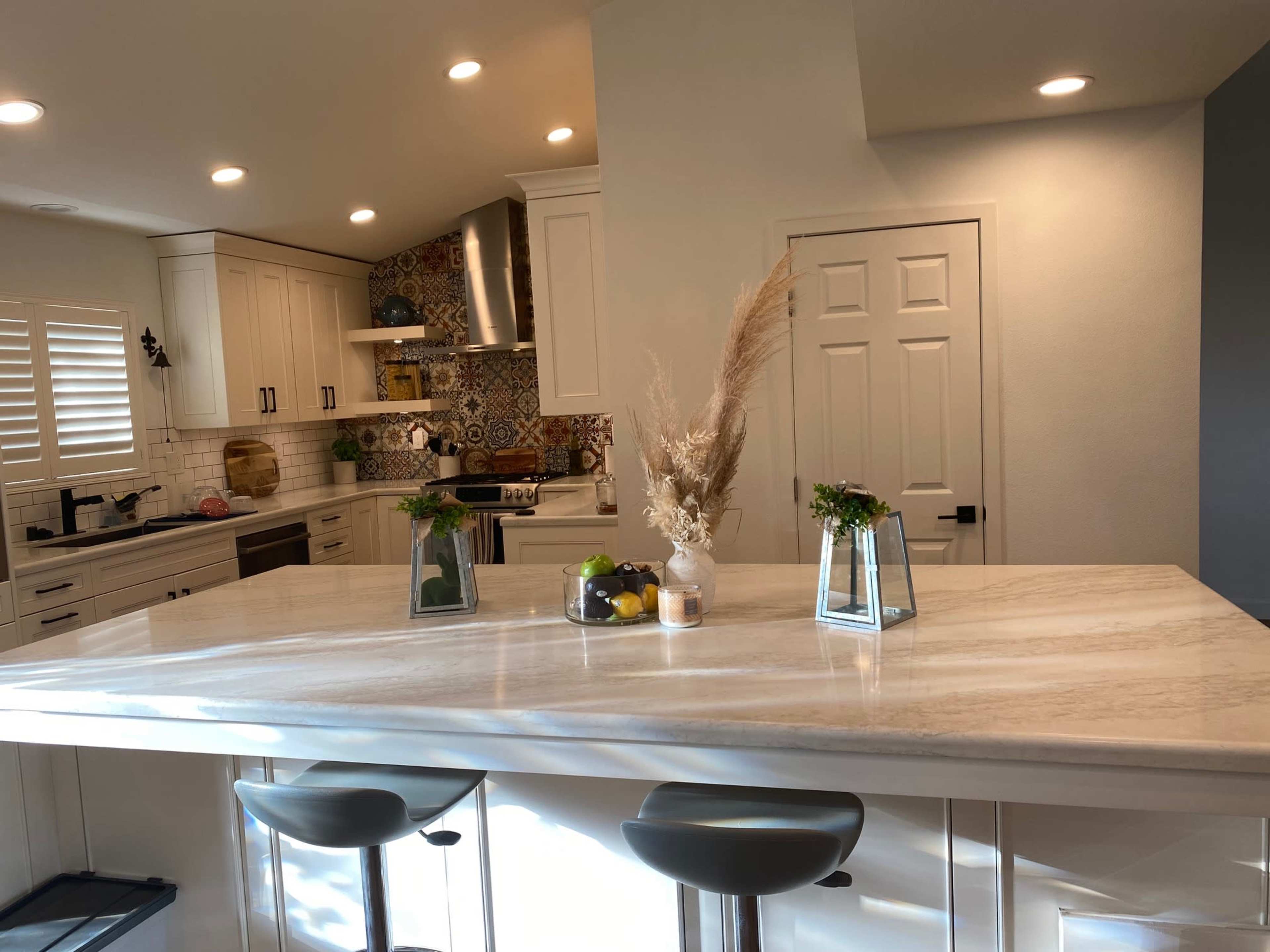 A modern kitchen features a large marble island with plants and fruit arrangements, white cabinets, and a decorative tiled backsplash.