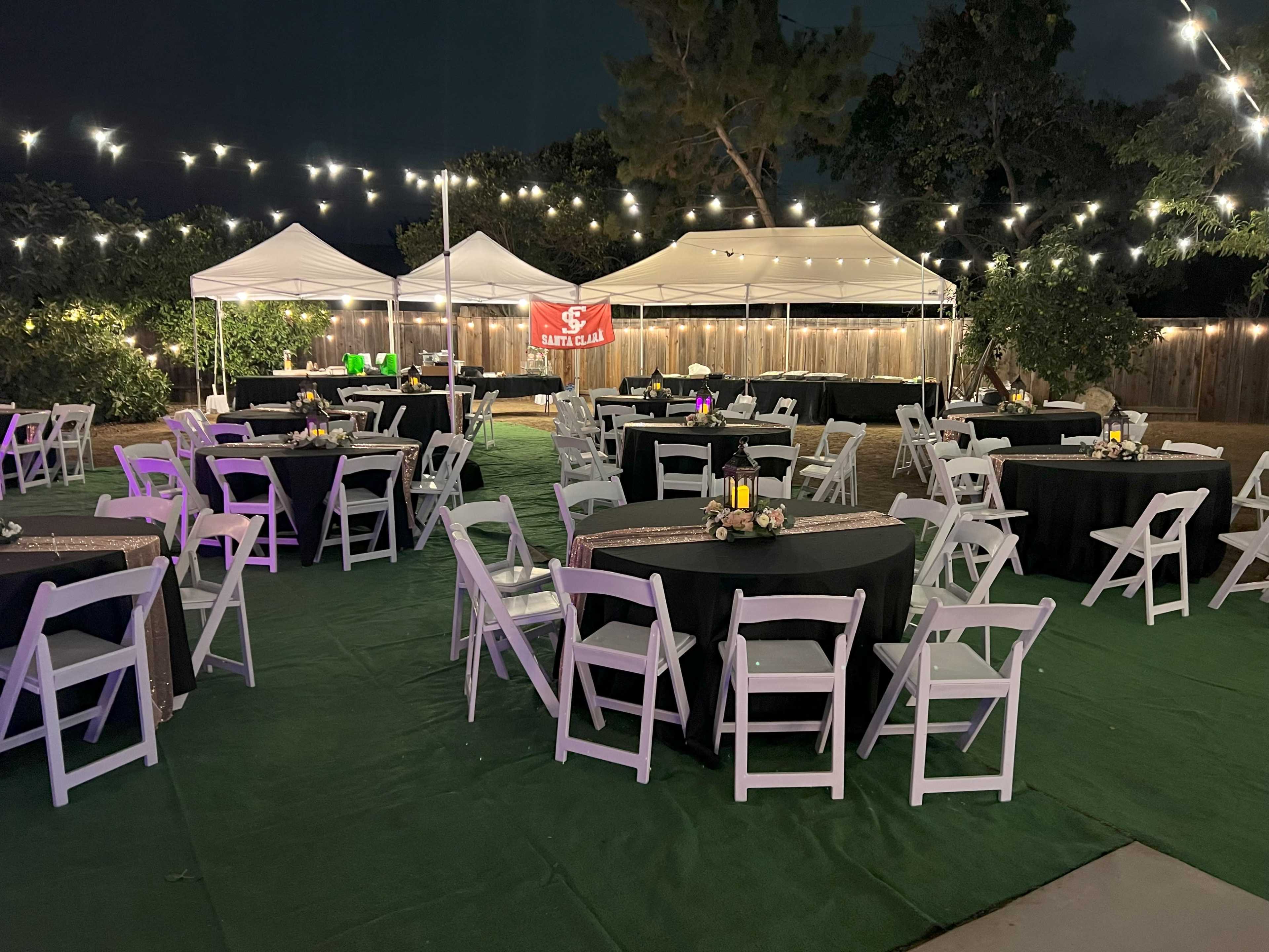The image shows a nighttime outdoor event space set up with multiple tables and tents, adorned with string lights overhead.