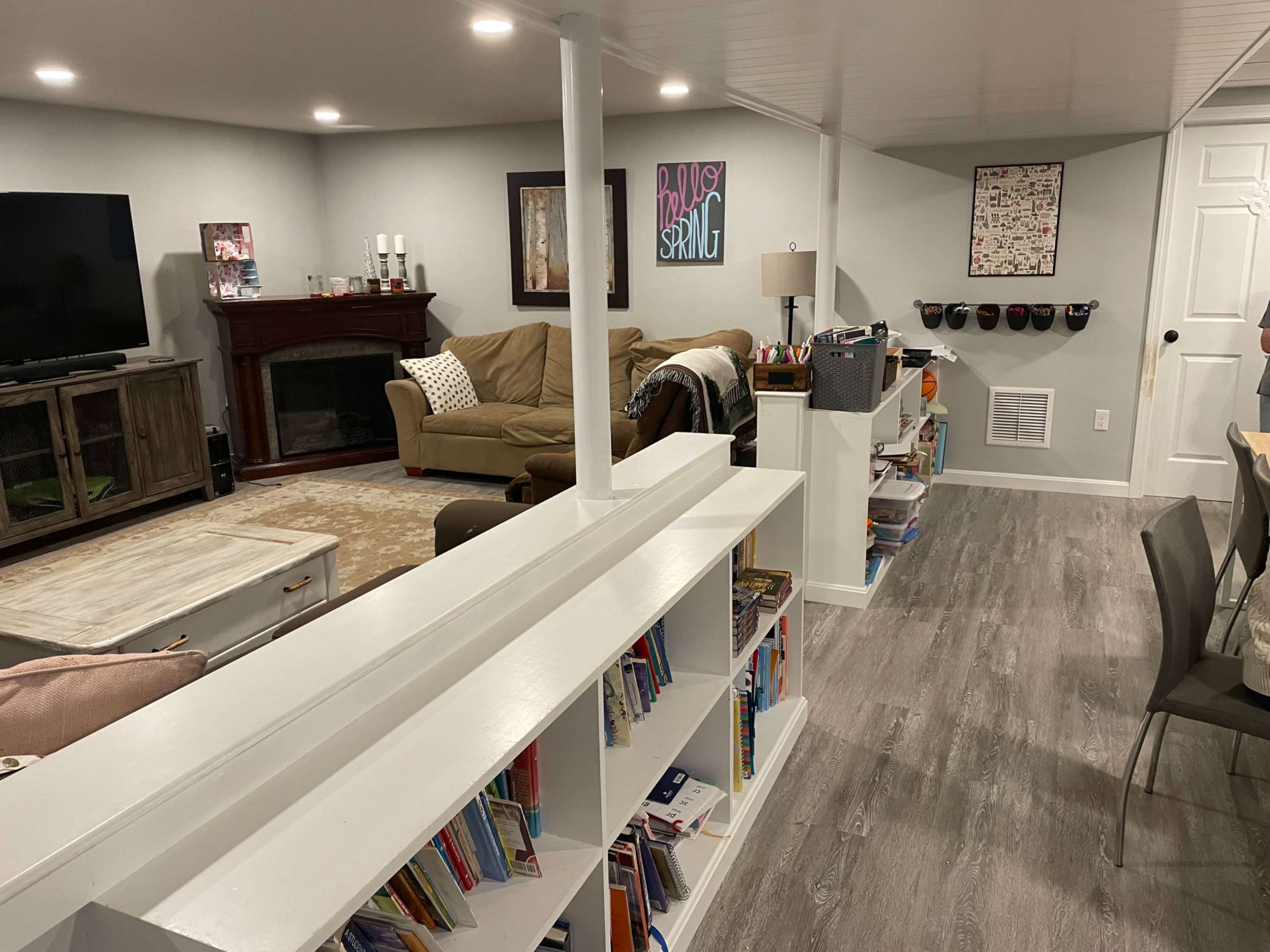 The image shows a finished basement with a seating area featuring a couch and television, a bookshelf filled with various books, and a small dining table with chairs.