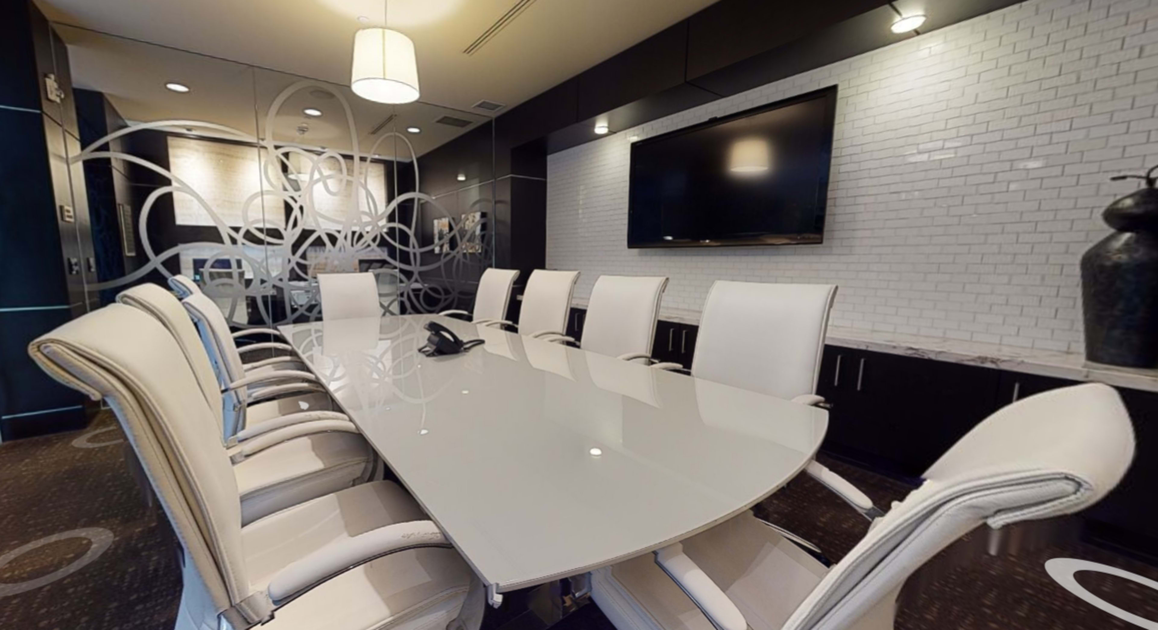A modern conference room features a large white table surrounded by white chairs, with a television on the wall and glass partitions.