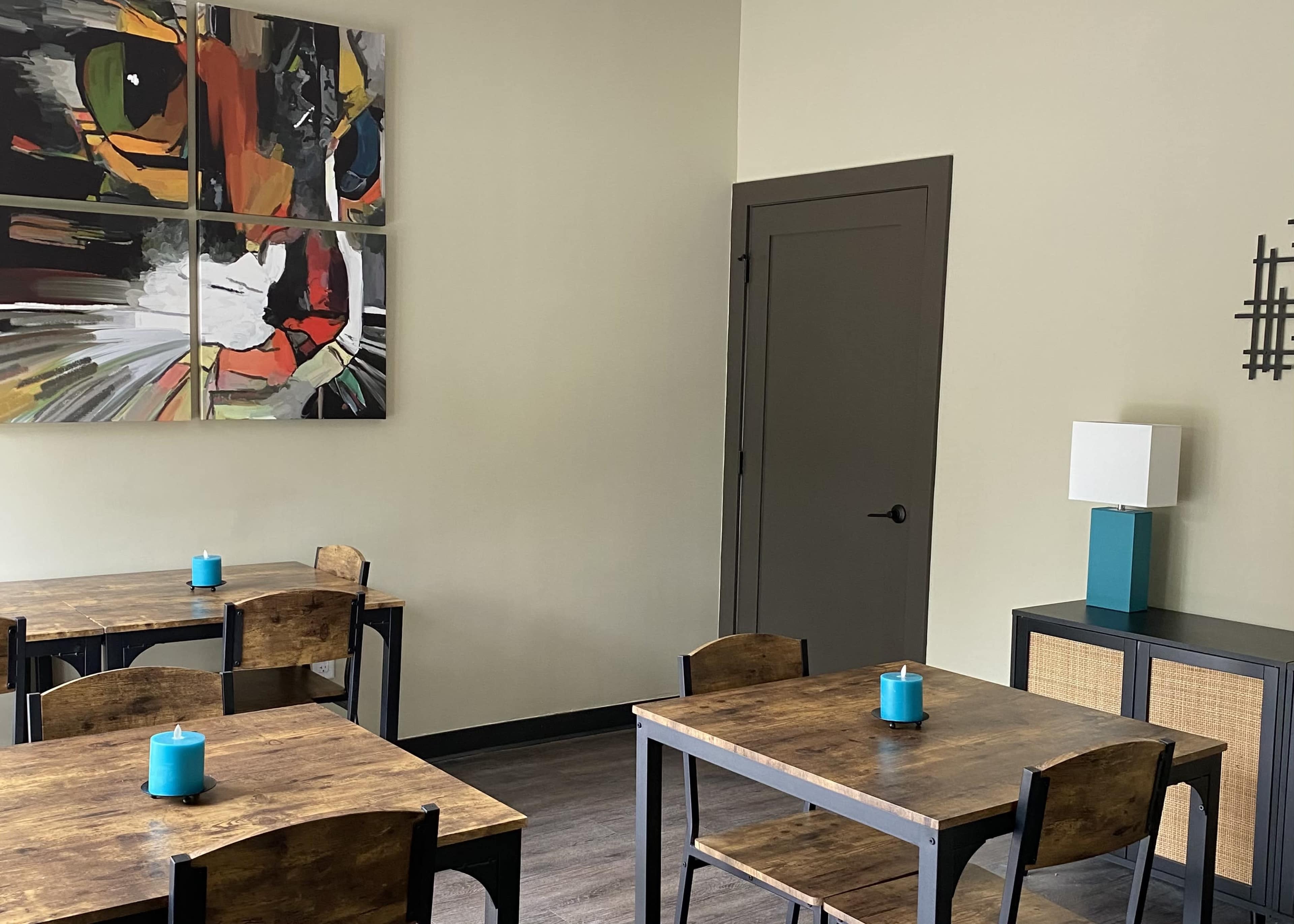 A contemporary dining area with wooden tables, teal candles on each table, and a large colorful cat painting on the wall.