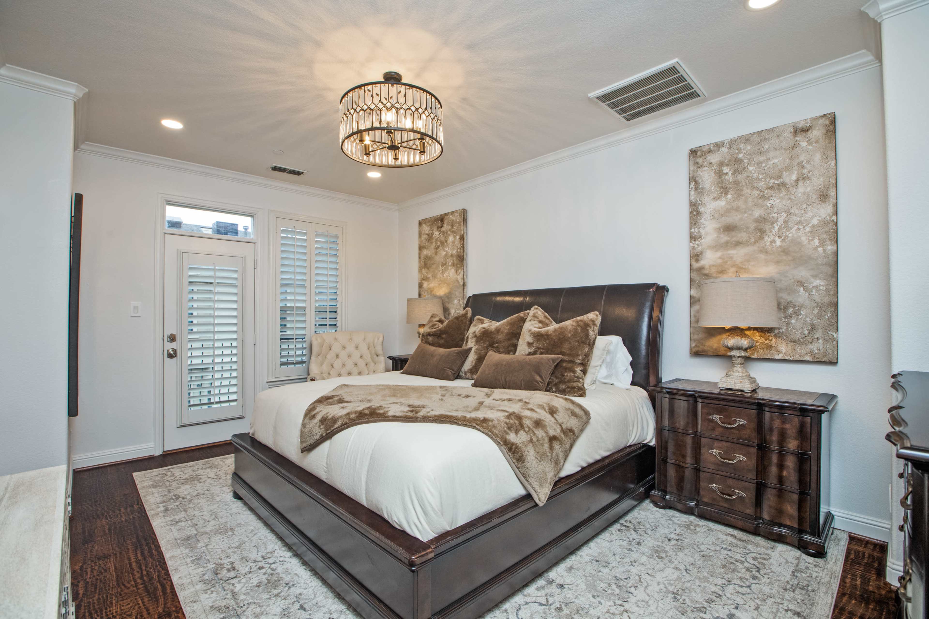 A modern bedroom features a large bed with decorative pillows, a chandelier, two artwork pieces on the walls, and a wooden dresser beside a door leading outside.