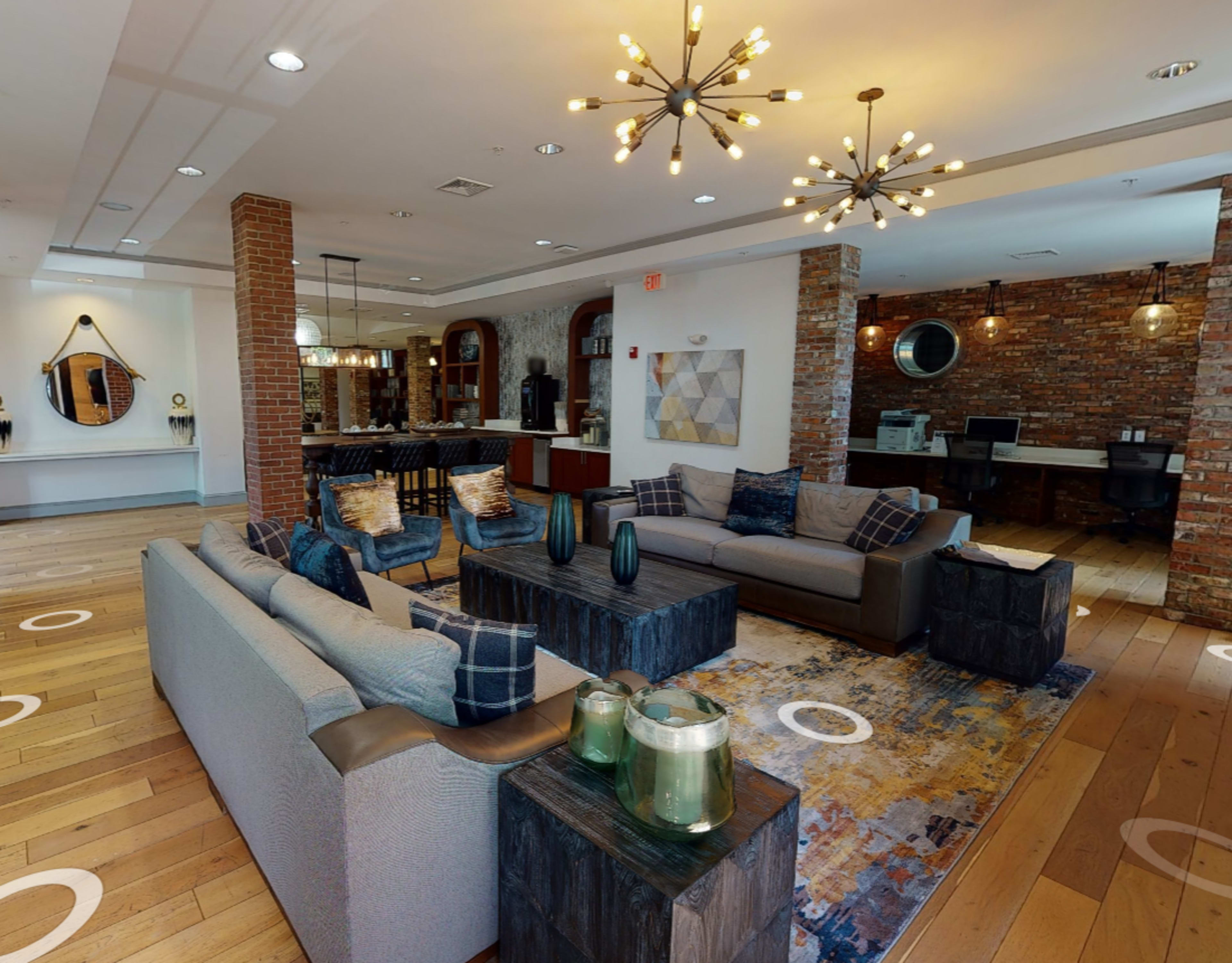A modern lounge area with two gray sofas, wooden tables, a decorative rug, and exposed brick walls, creating a welcoming space.