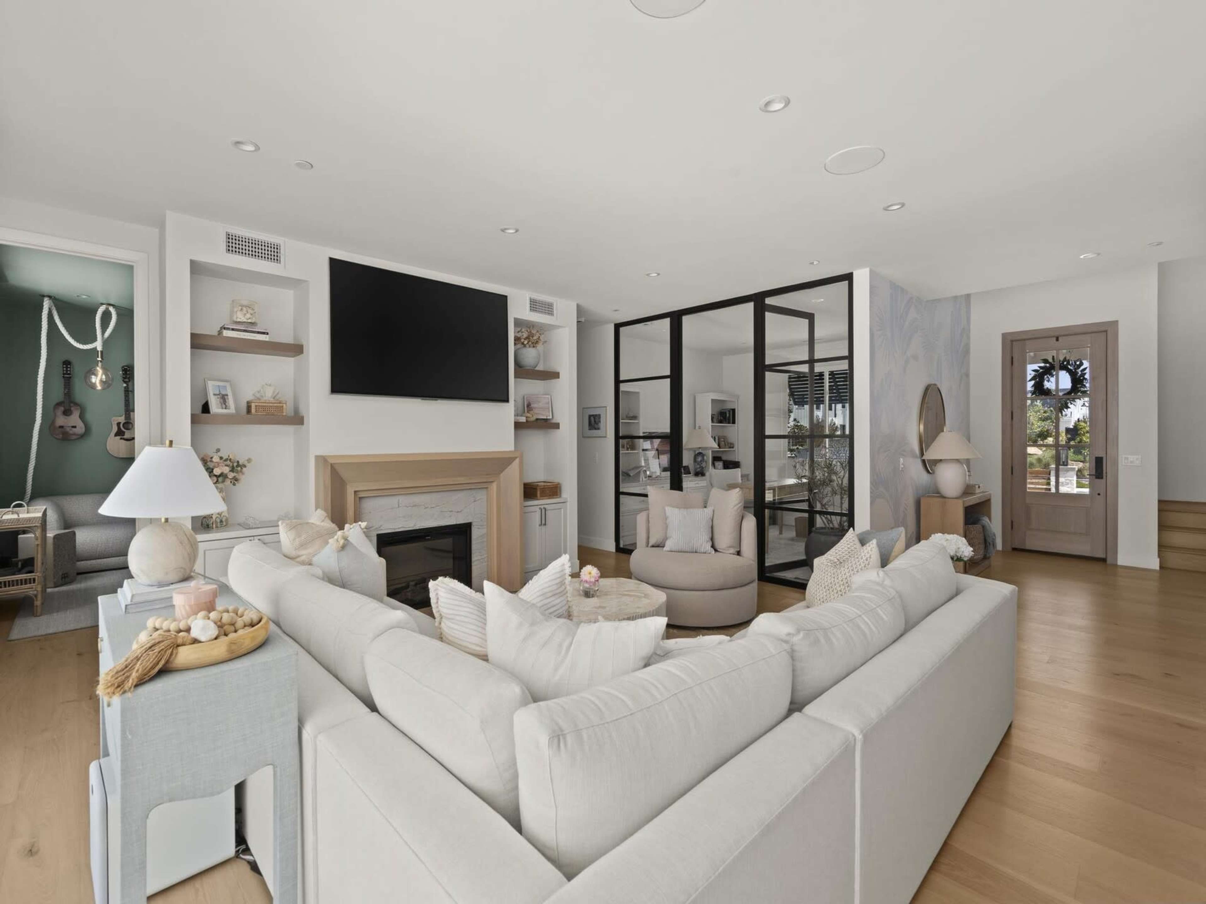 The image depicts a modern living room featuring a large sectional sofa, a wall-mounted television, a glass partition leading to another room, and wooden flooring.