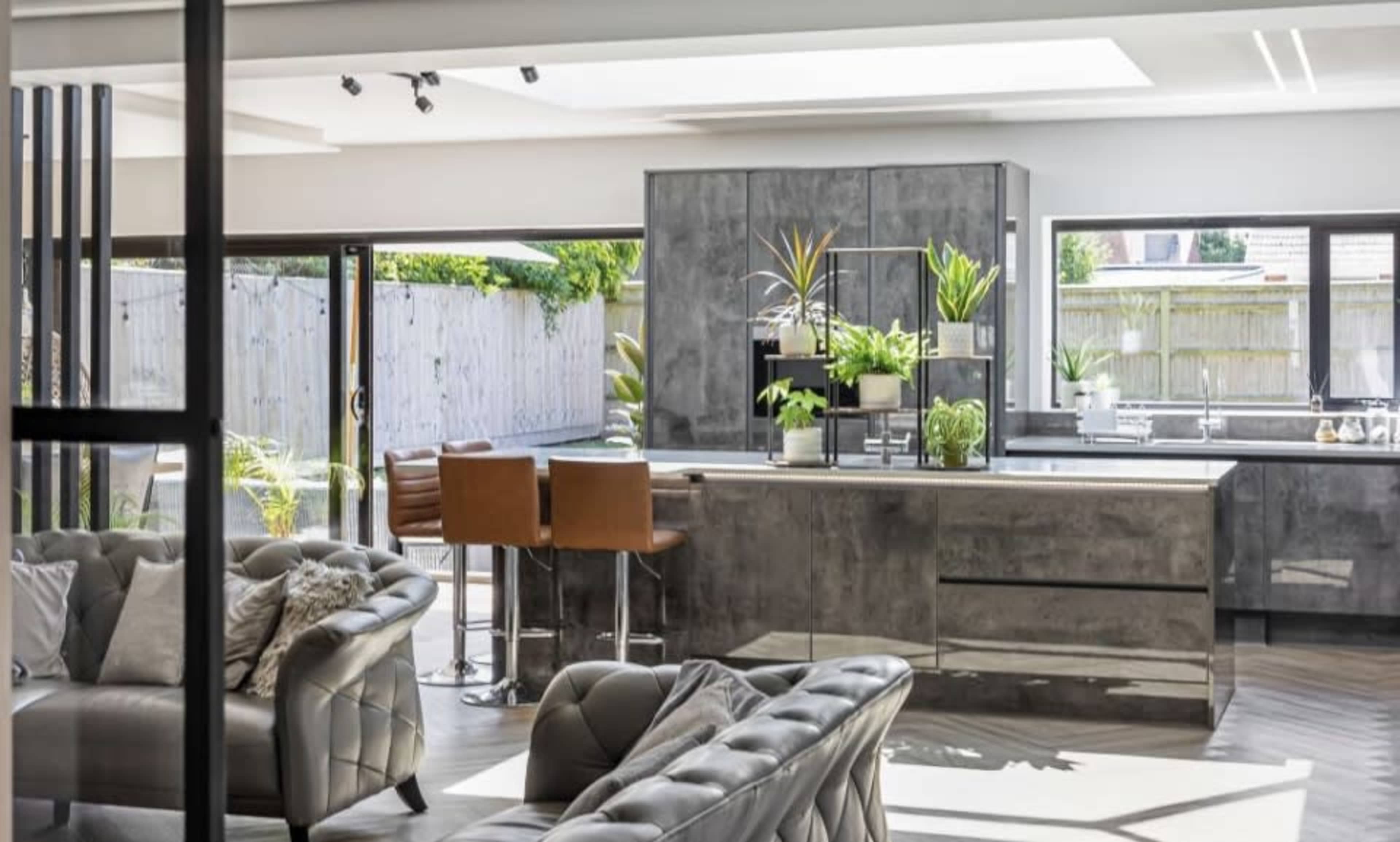 The image shows a modern kitchen and living area with sleek cabinetry, a central island, and large windows that open to a backyard garden.
