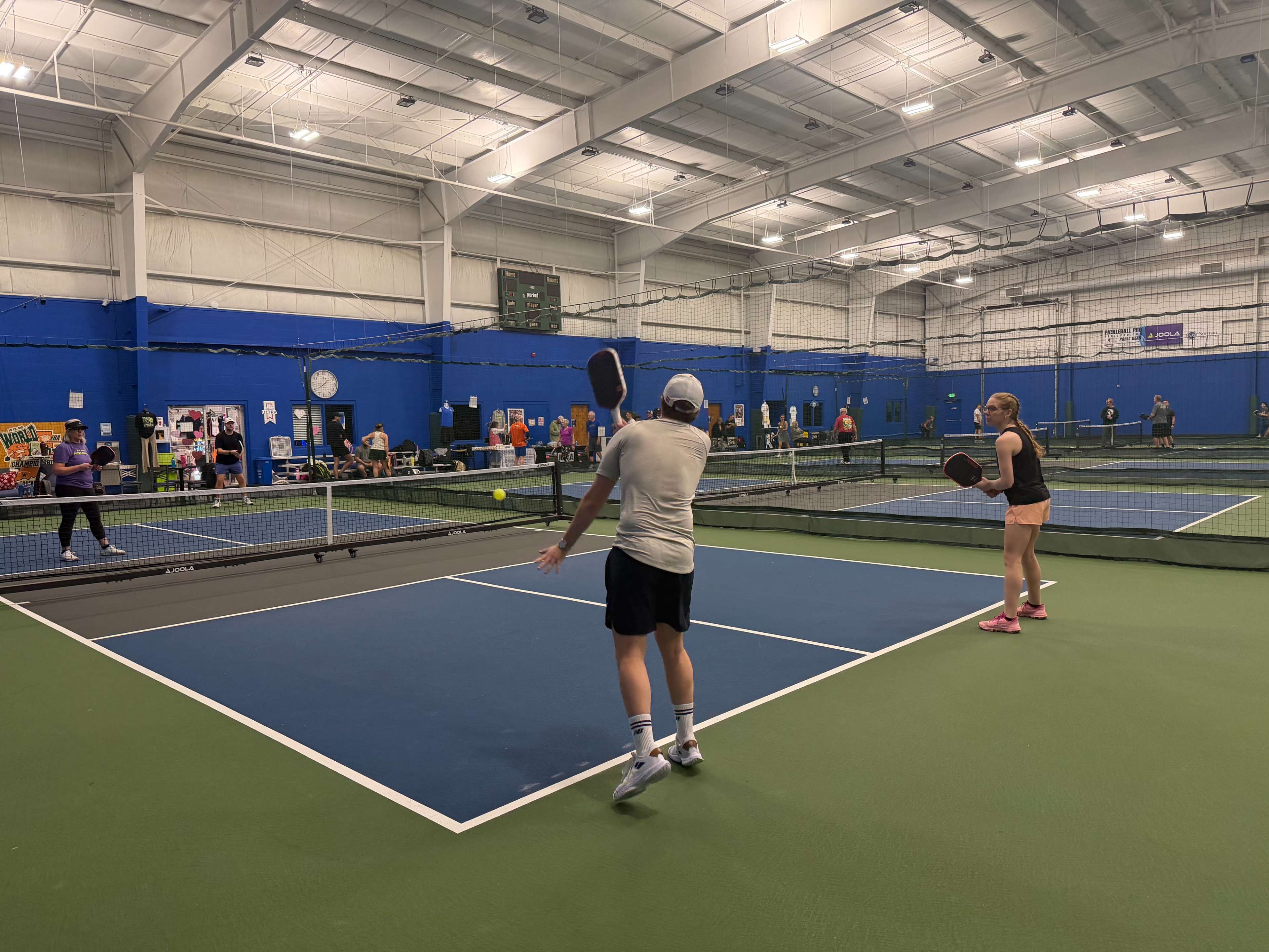 Two tennis players are actively engaged in a match inside a well-lit indoor tennis facility, with multiple courts visible in the background.