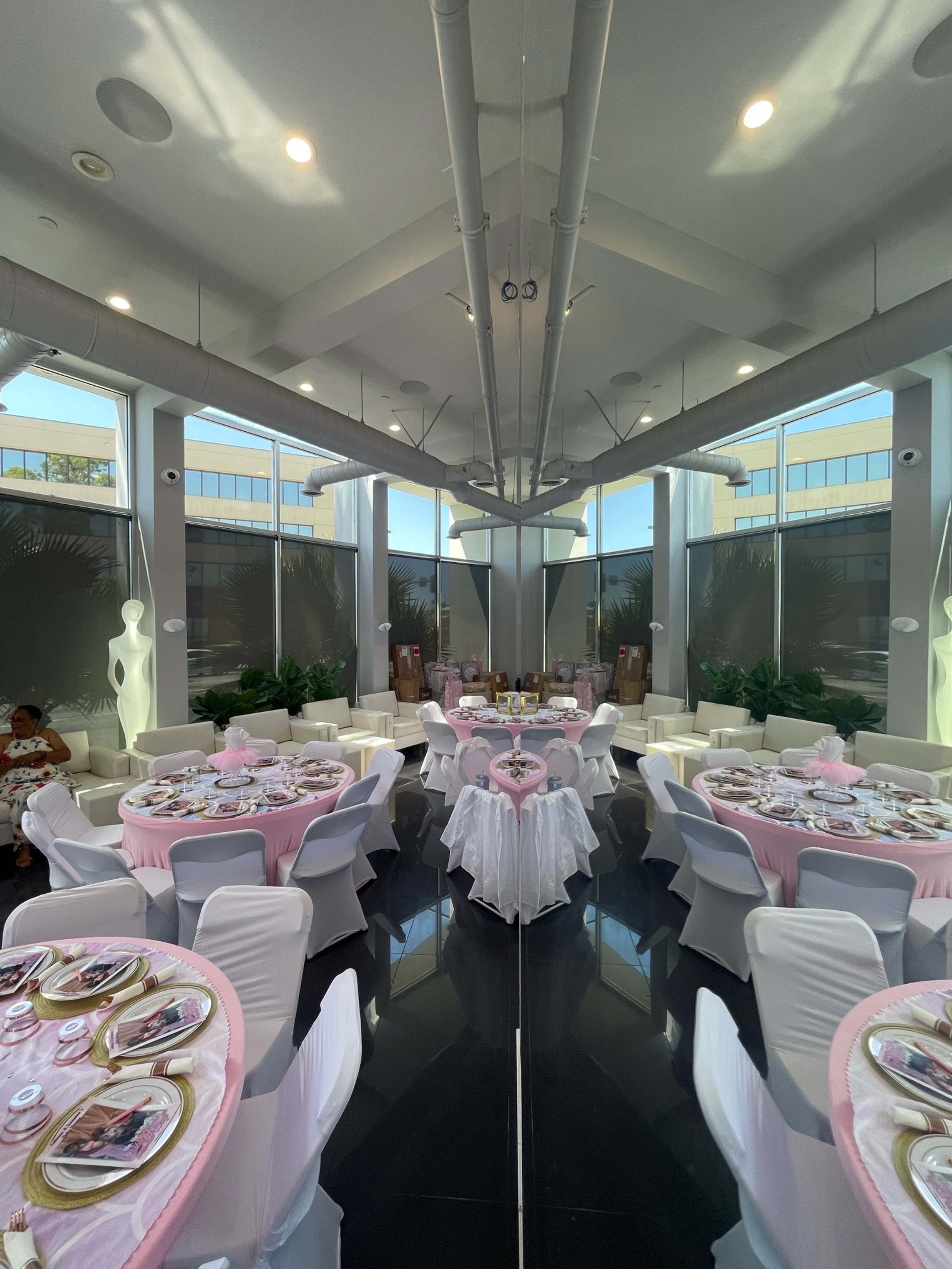 The image shows a neatly arranged dining area with round tables covered in pink tablecloths, set for a formal event, illuminated by natural light coming through large windows.