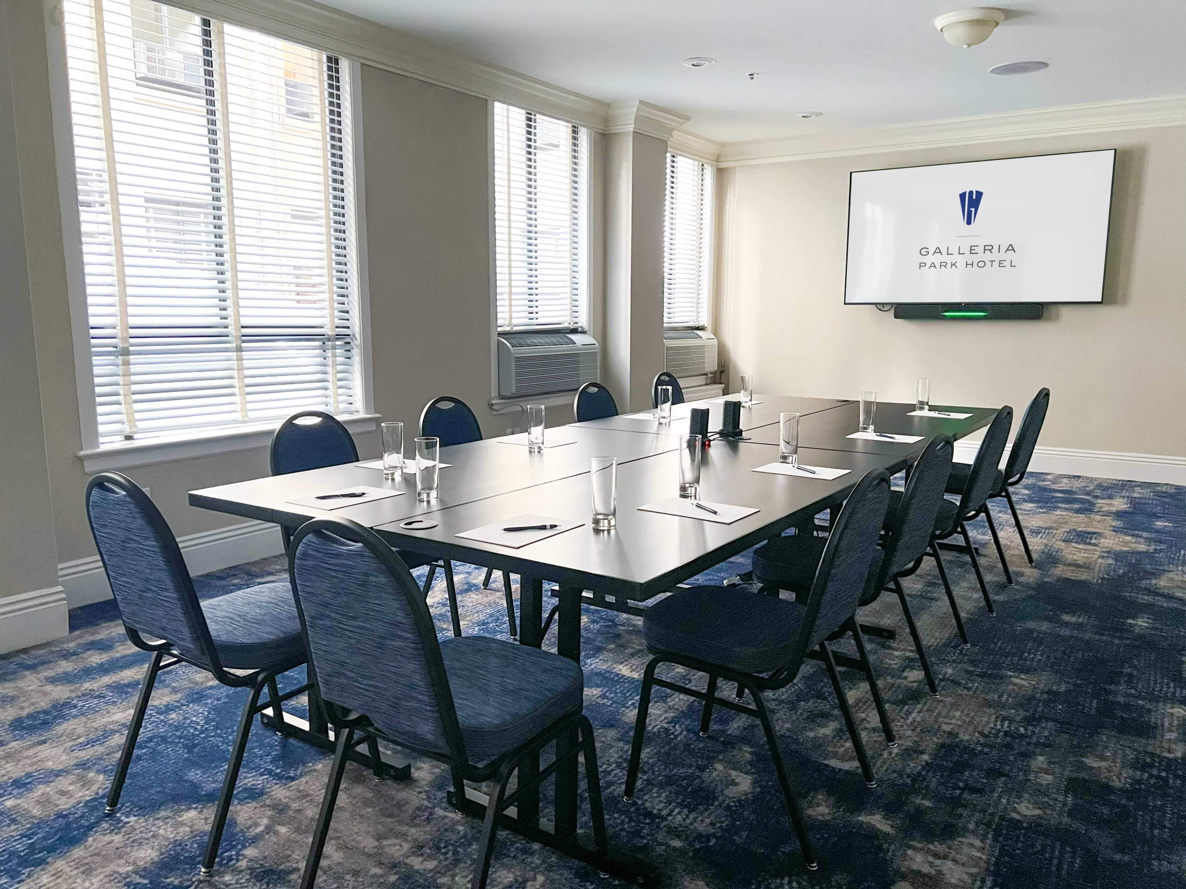 A meeting room is set with a long table, chairs, and presentation screen, with natural light coming through the windows.