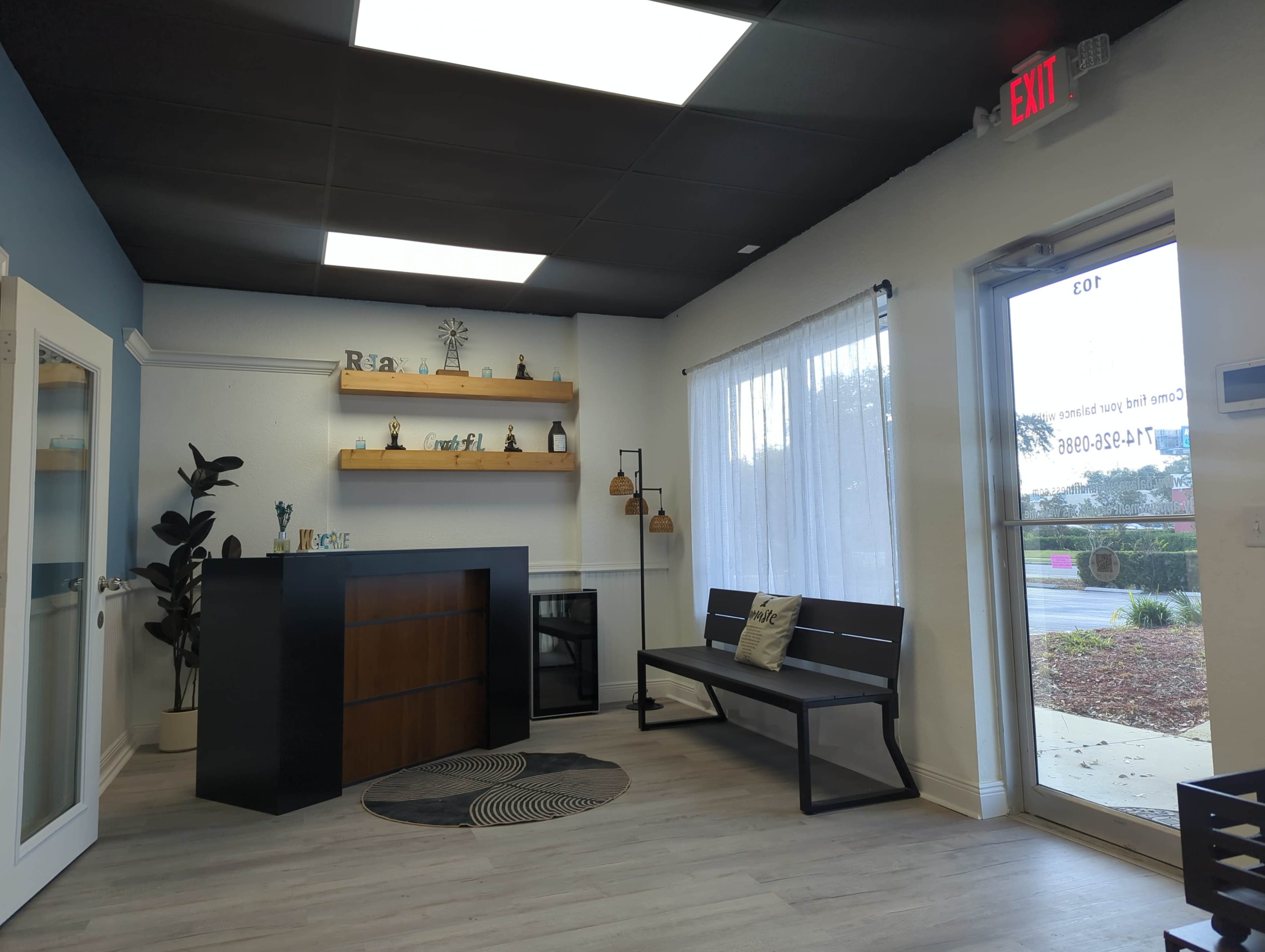 The image shows a modern reception area with a dark wood counter, a bench, and decorative shelves, illuminated by natural light from large windows.