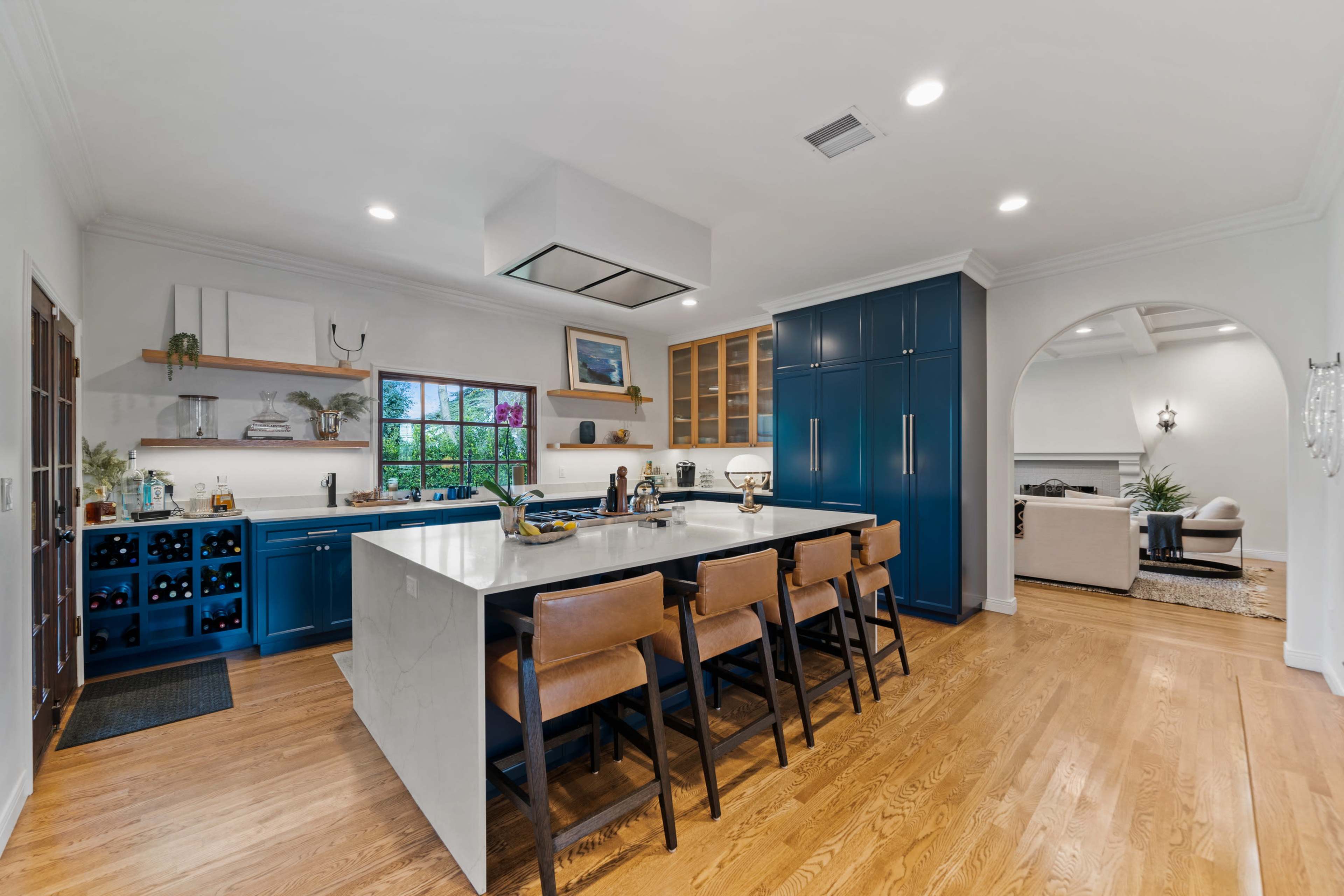 A modern kitchen features blue cabinetry, a large island with seating, and a view of an adjoining living area.