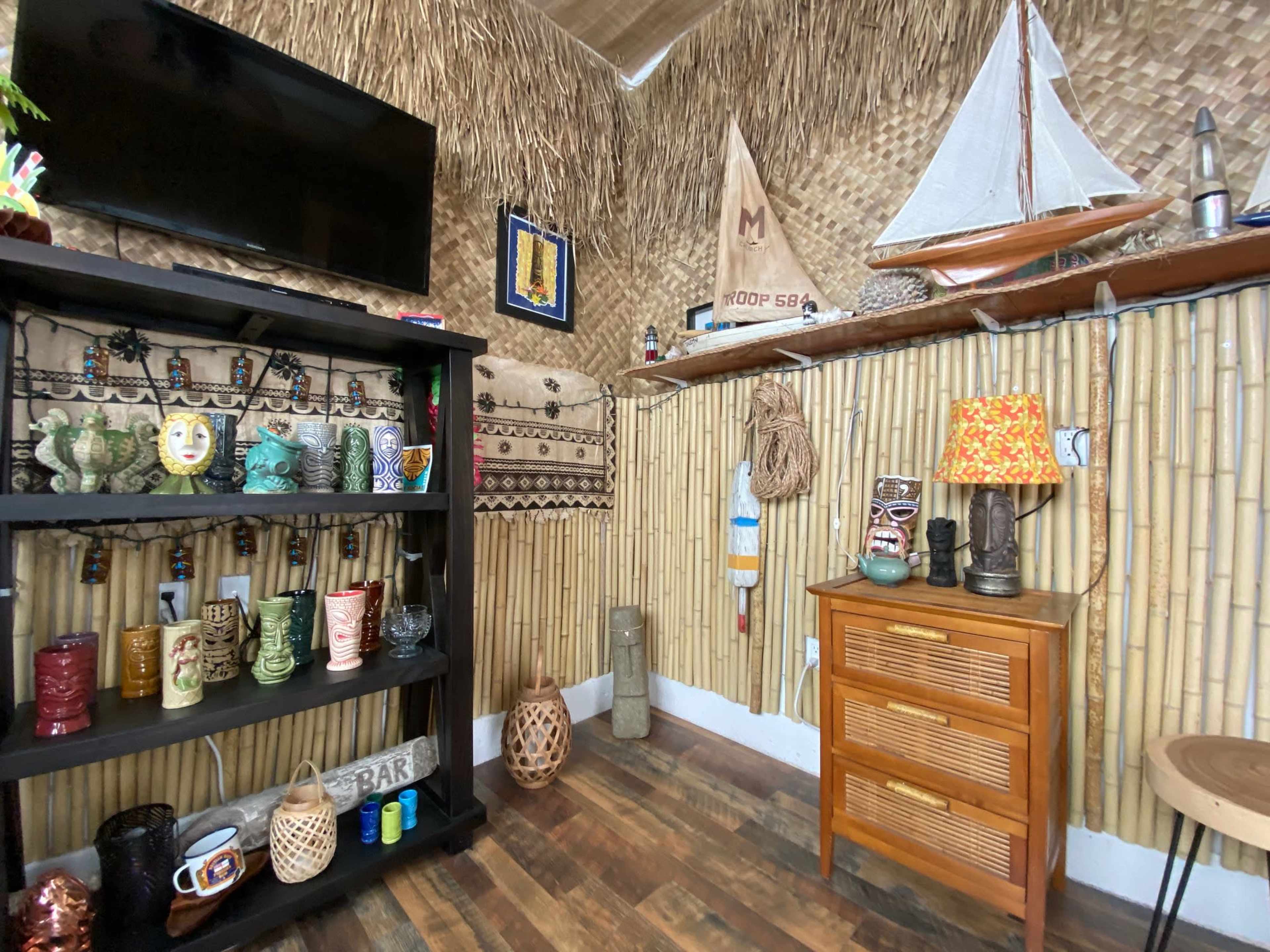 A tropical-themed interior featuring a bamboo wall, decorative items including a model sailboat, a wooden cabinet with various colorful mugs, and a lamp on a side table.