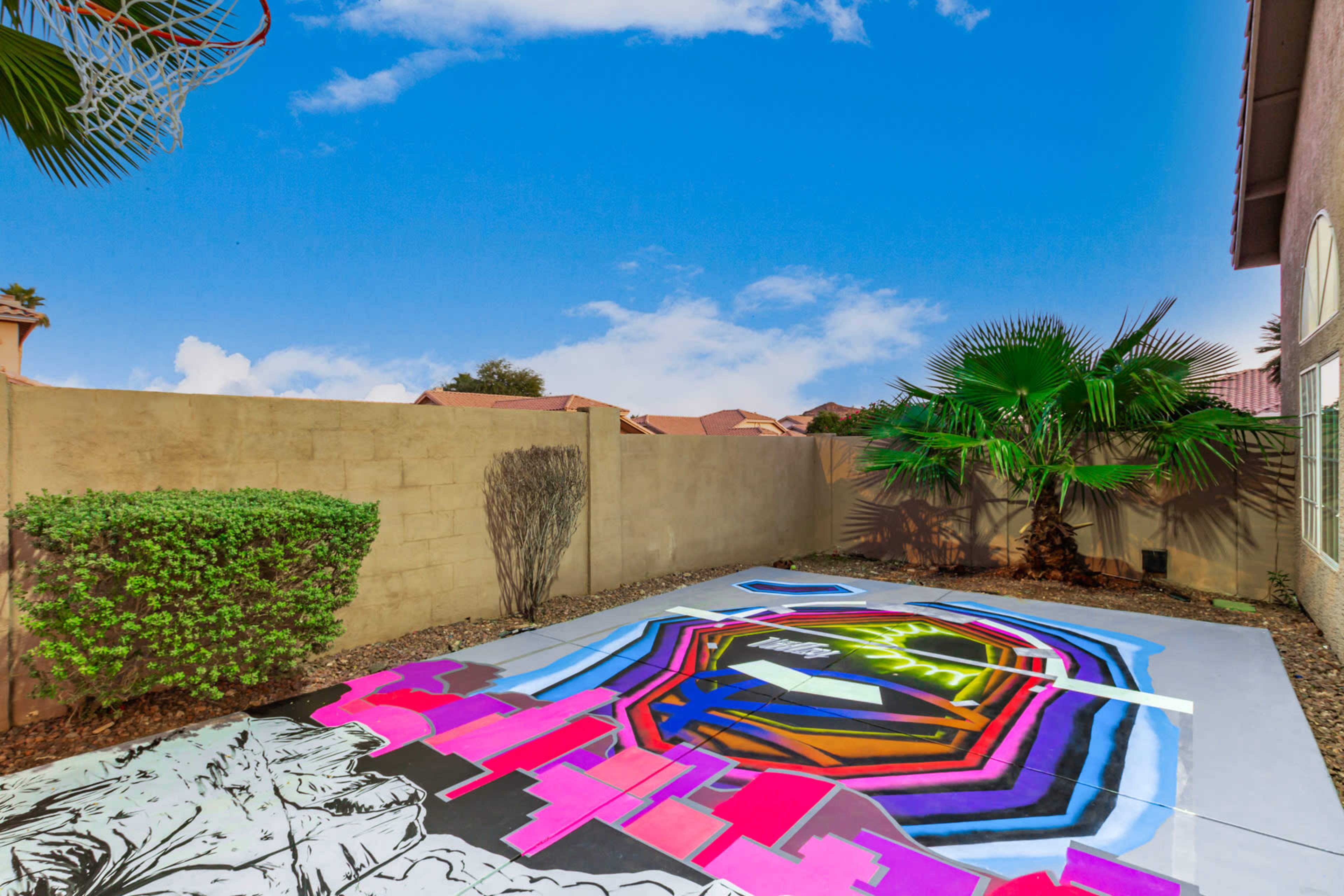 A brightly painted outdoor basketball court featuring geometric designs and vibrant colors is surrounded by a concrete wall and tropical plants.