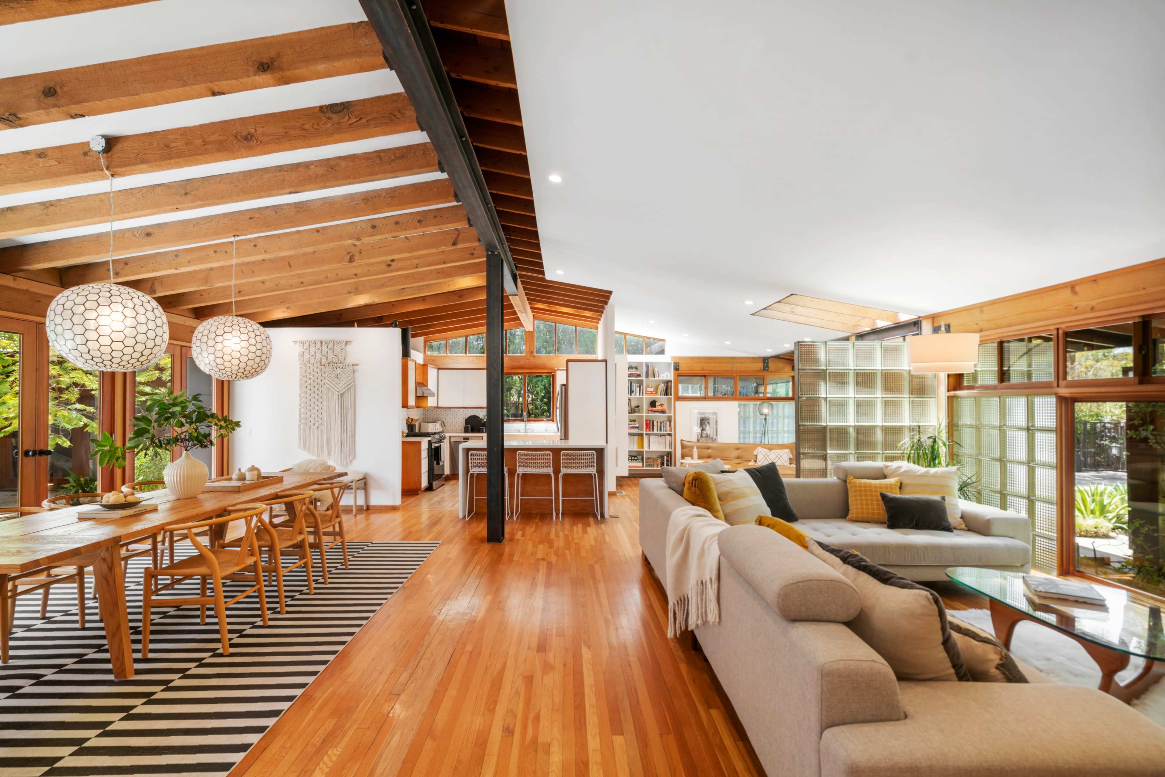 An open-concept living area with wooden beams, a dining table, and a comfortable sofa, surrounded by large windows allowing natural light.
