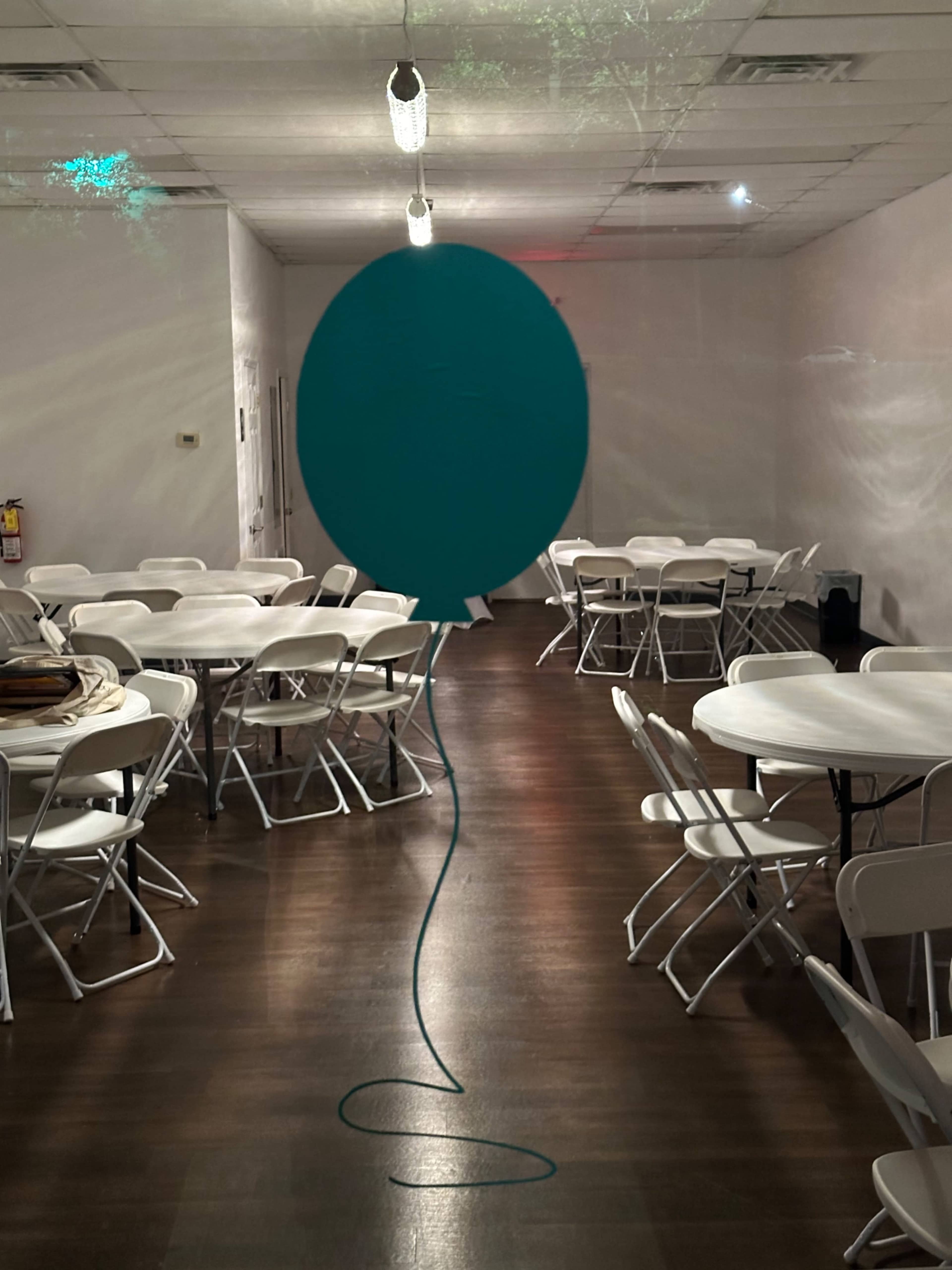 A large teal balloon with a long string is centered in a spacious, empty room filled with round tables and chairs.
